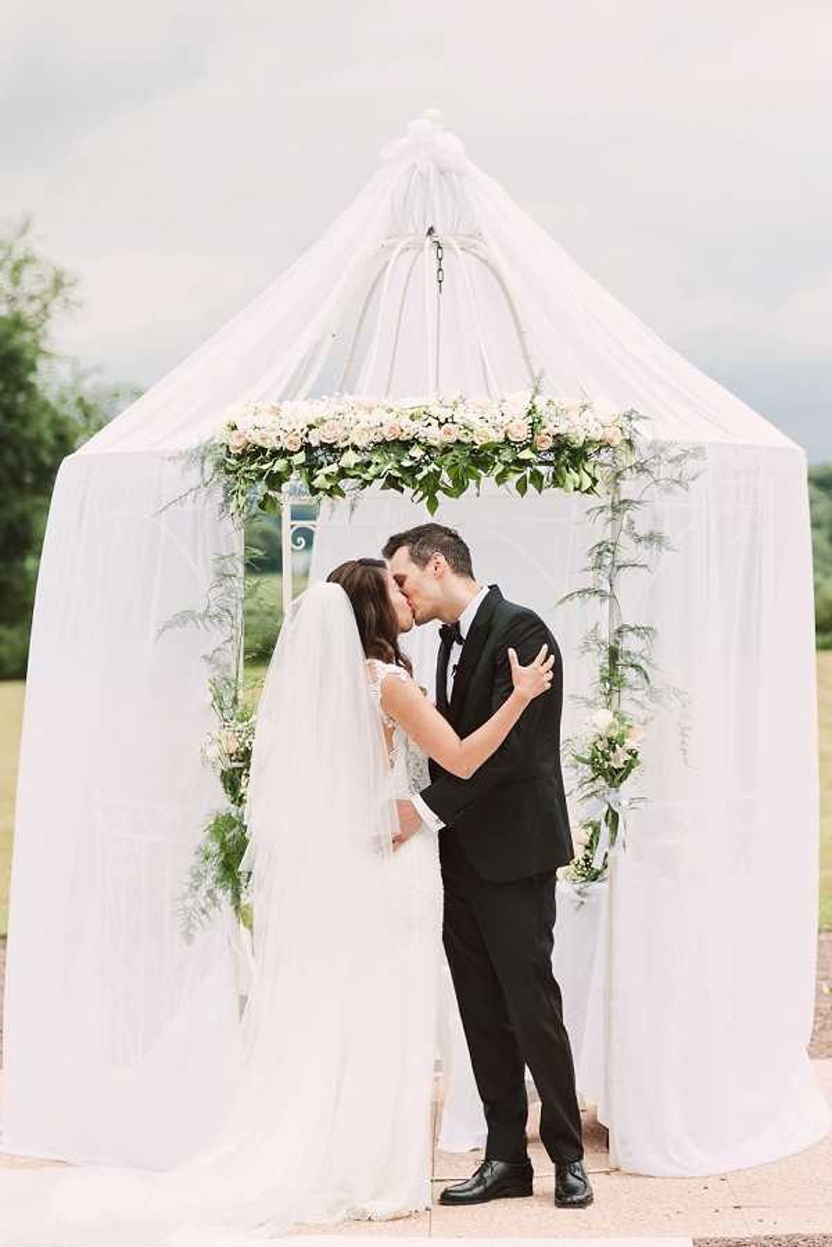 A bride and groom share a kiss during an outdoor ceremony beneath a white draped gazebo structure with a tent-like canopy. The gazebo is decorated with a floral garland of blush and ivory roses with lush greenery along its upper interior frame, and additional greenery and small florals accent the side posts. The bride wears a lace-detailed white gown with a long cathedral-length veil, while the groom wears a classic black tuxedo with a bow tie. The portrait is taken as a medium-wide shot with the couple centered against the white draped backdrop, set within an open outdoor garden or estate grounds setting with a classic, formal styling aesthetic.
