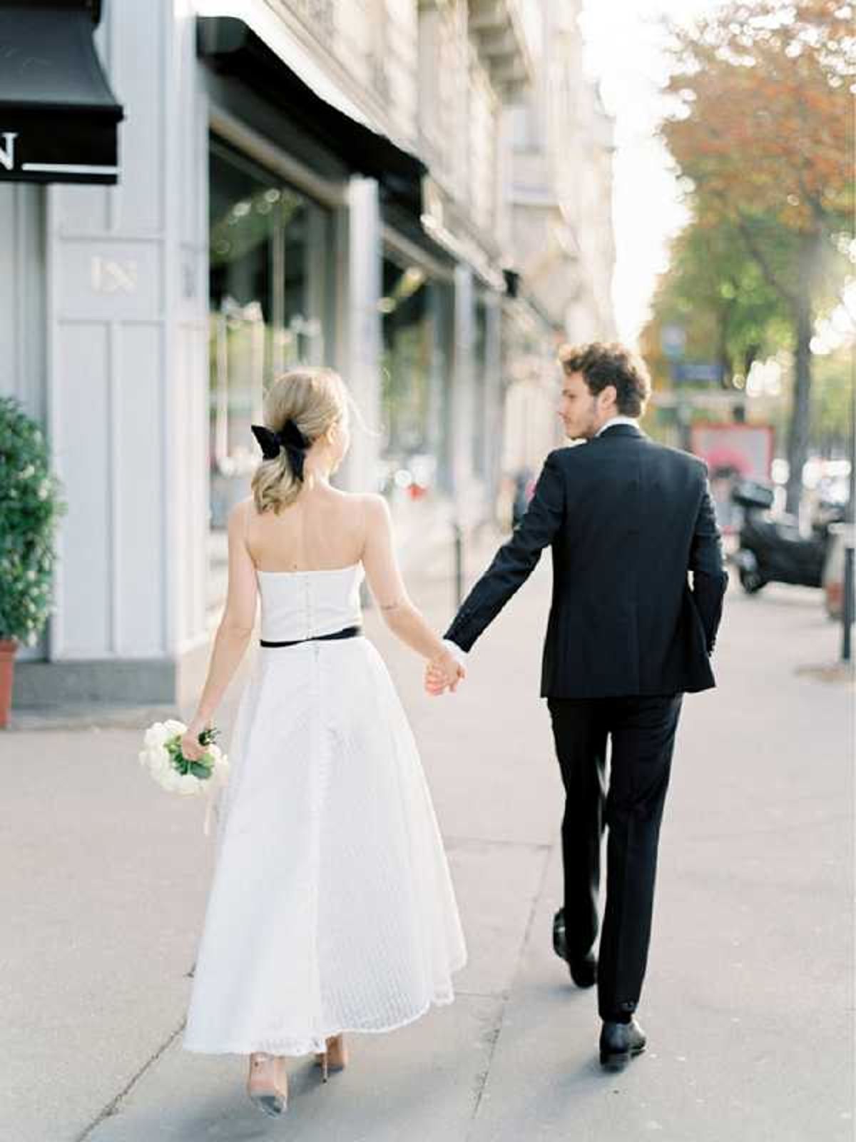 A couple walks hand-in-hand along a Parisian city sidewalk in this portrait-style shot taken from behind. The bride wears a strapless, tea-length white dress with a thin black belt at the waist, accessorized with a large black bow in her low blonde ponytail, and carries a small bouquet of white blooms; the groom is dressed in a classic black suit with black dress shoes. The composition is a mid-distance shot with the couple centered against a boulevard lined with Haussmann-style building facades featuring black awnings, shot in warm golden-hour light. The overall styling is clean and minimal with a monochromatic black-and-white color theme.