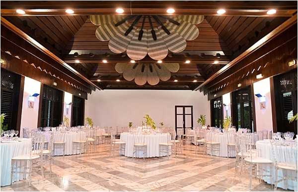 Wide shot of an indoor wedding reception venue set up for a seated dinner, featuring round tables dressed in white linens and surrounded by clear ghost/chiavari chairs. The room has a distinctive dark wood ceiling with exposed beams and a large decorative ceiling installation made of overlapping petal-shaped panels in a striped black-and-white pattern. The marble or polished tile floor has a geometric pattern and serves as the dance floor in the center. Table centerpieces appear minimal with small greenery accents. The overall decor palette is white and clear with dark wood architectural details, giving a clean, tropical-colonial aesthetic. Louvered dark wood shutters line the walls, and recessed ceiling lighting illuminates the space. Potential venue feature image.