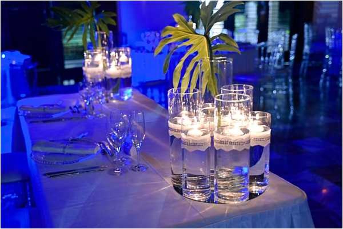 A close-up detail shot of a wedding reception table set in an indoor venue with dramatic blue uplighting. The centerpiece features tall clear cylinder vases with floating candles and submerged white floral elements, grouped together alongside a taller vase holding tropical monstera leaves. The white linen tablecloth is set with wine glasses, silverware, and white folded napkins on white charger plates accented with small beaded details. The decor palette is modern and minimal, combining white, clear glass, and tropical greenery against the vivid blue ambient lighting that fills the background room.