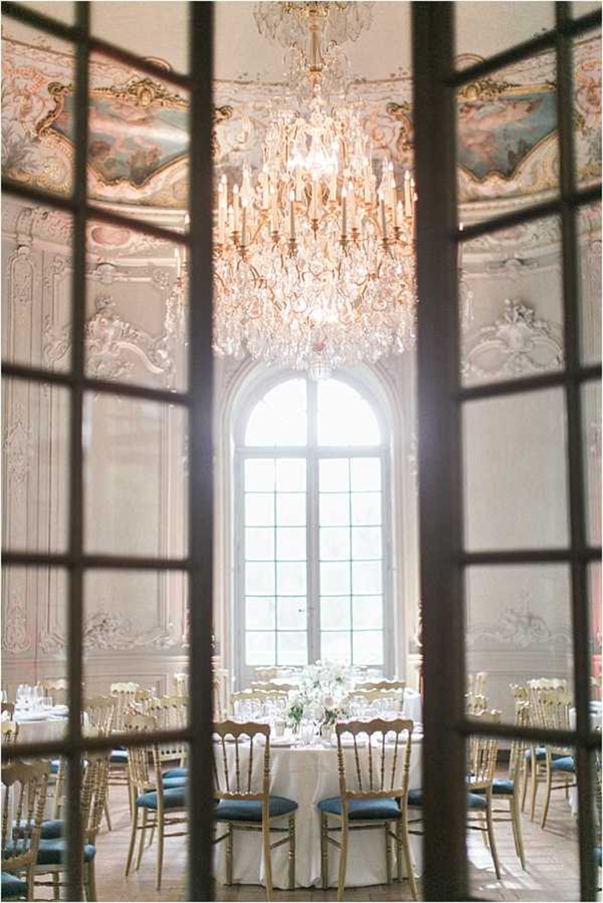 A wide shot of an indoor wedding reception room photographed through the panes of tall French doors, which frame the composition in the foreground. The ballroom features ornate Baroque-style plasterwork on the walls and ceiling, painted ceiling medallions in soft blue and gold tones, and a large crystal and gilt candelabra chandelier hanging at center. A tall arched window floods the back of the room with natural light. Round dining tables are set with white linen cloths, gold Chiavari chairs with teal/slate blue cushions, glassware, and low centerpieces of white and blush flowers. No people are present; the room is set up and awaiting guests. The overall decor palette is white, gold, and teal against the cream and gilt architecture. Potential venue feature image.