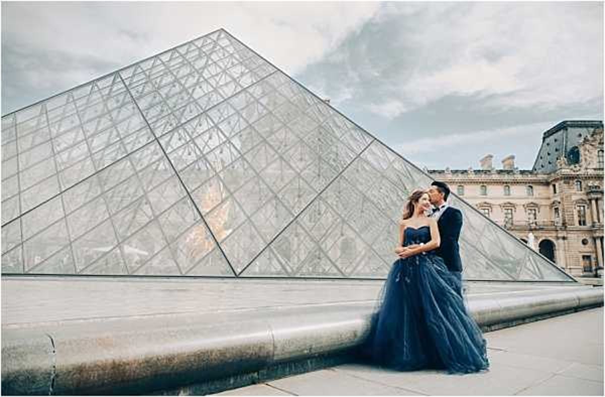 A couple poses for a pre-wedding or engagement portrait in front of the Louvre Pyramid in Paris, with the historic Louvre Palace building visible in the background. The bride wears a strapless navy blue tulle ballgown, and the groom is dressed in a matching navy blue suit, creating a coordinated color scheme. The two stand close together in an intimate embrace, leaning toward each other, with the glass-and-steel pyramid filling most of the frame behind them. This is a wide environmental portrait shot, taken at ground level, with overcast natural light.