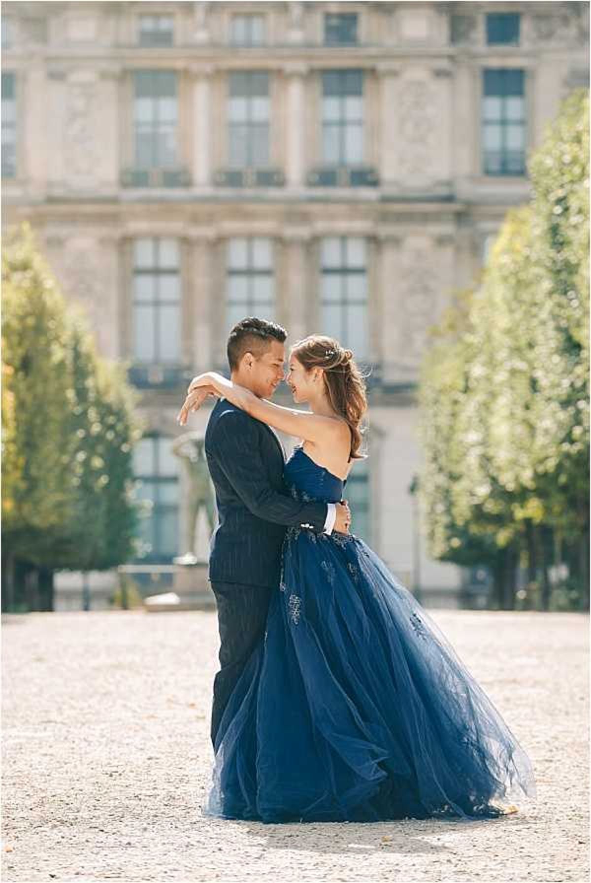 A couple poses together in an outdoor portrait session on a gravel esplanade in front of a large classical French palace or institutional building, consistent with Paris architecture. The bride wears a strapless navy blue tulle ballgown with beaded or embroidered detailing, and her hair is styled in a loose updo with a small hair accessory. The groom wears a dark navy pinstripe suit. They are facing each other closely, smiling, with her arms around his neck and his hands at her waist. The composition is a medium portrait shot with the ornate building and formally trimmed trees softly blurred in the background, creating strong depth of field. The styling leans toward a classic, formal aesthetic with the coordinating navy color palette.