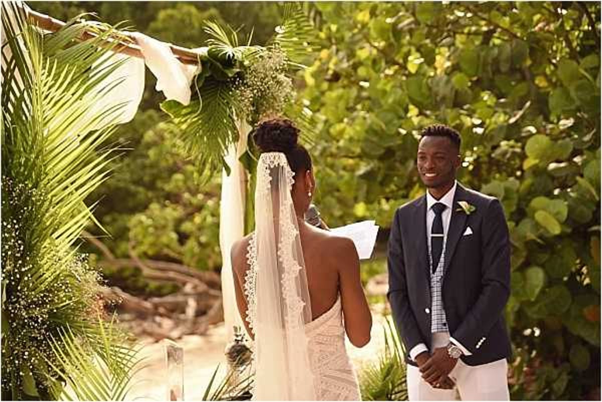 An outdoor beach wedding ceremony showing the bride and groom at the altar, with the bride seen from behind reading her vows and the groom facing the camera smiling. The bride wears a backless lace gown with a long lace-trimmed veil and her hair in an updo, while the groom wears a navy suit jacket with white trousers, a patterned tie, and a white boutonniere. The ceremony arch is decorated with tropical greenery including palm fronds and baby's breath with white fabric ribbon accents. The composition is a medium portrait shot capturing both figures at the altar with lush tropical foliage in the background.