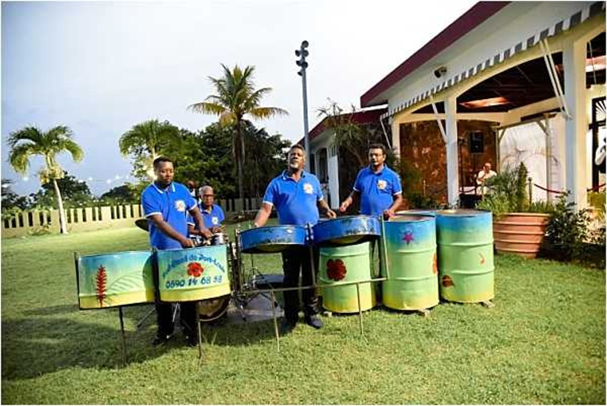 A steel drum band of four musicians performs outdoors on a lawn during what appears to be a wedding cocktail hour or reception. The musicians are dressed in matching blue polo shirts and are playing colorful painted steel pan drums — the barrels feature tropical-themed artwork in yellow, green, and red tones, with a phone number and text visible on one drum. The setting is an outdoor garden area in front of a low-rise venue building with a covered terrace, with palm trees visible in the background. The shot is a wide, candid performance photo taken at ground level during late afternoon or early evening light.