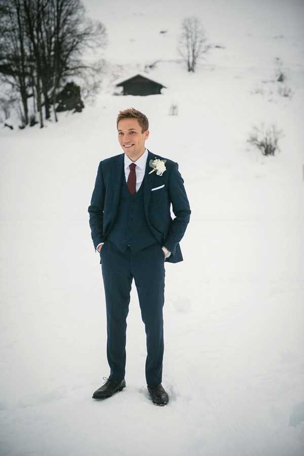 A groom portrait taken outdoors in a snowy winter mountain setting, with a wooden chalet visible in the background. The groom stands with hands in his pockets, wearing a navy three-piece suit with a burgundy tie, white pocket square, and a white floral boutonniere. He is smiling directly at the camera in a full-length portrait composition.