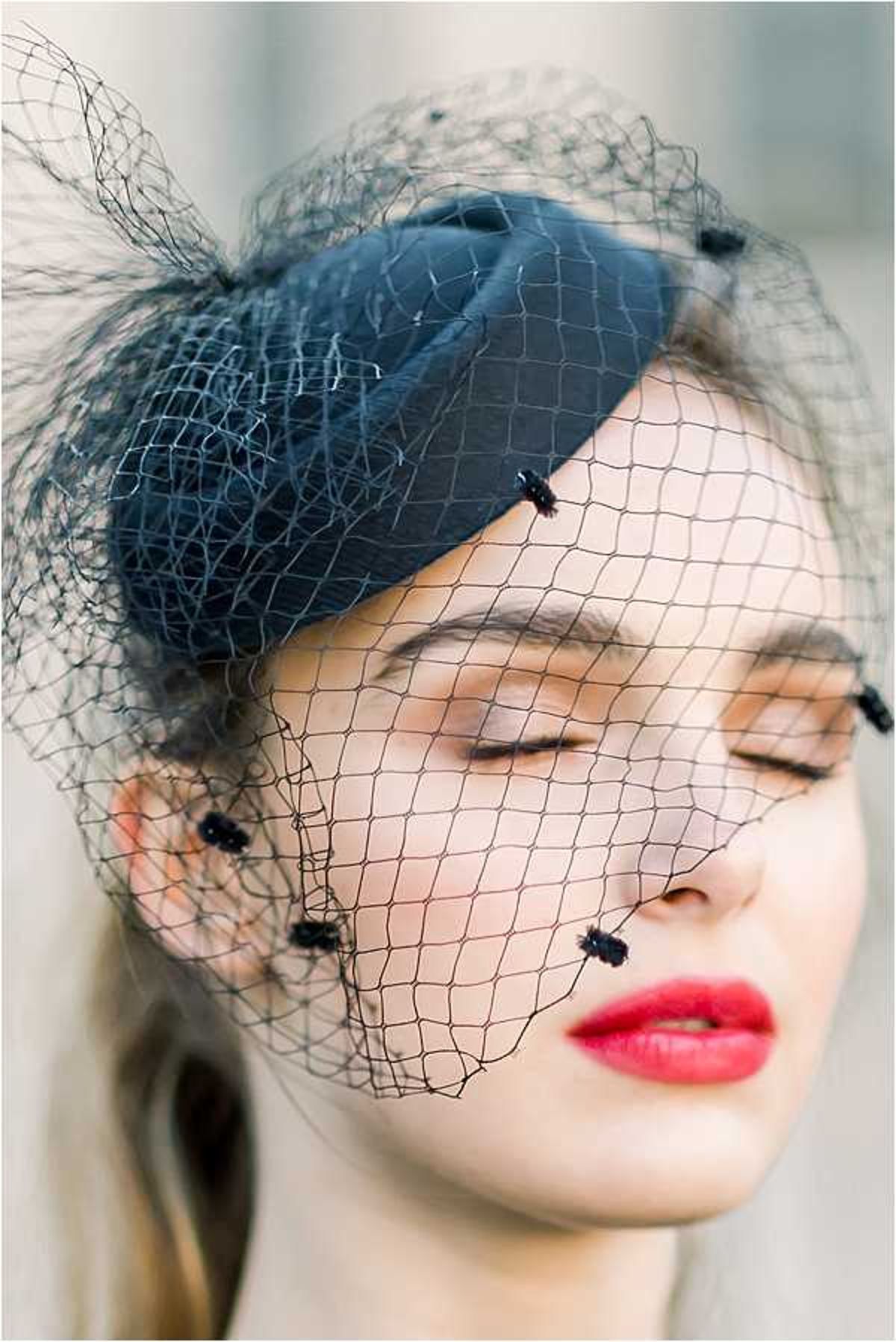 A close-up portrait of a woman wearing a navy blue satin fascinator with a matching navy birdcage veil adorned with small dark velvet or beaded knot details at the intersections. The subject has her eyes closed and wears a bold red lip, with blonde hair pulled back. The styling is vintage-inspired with a 1940s-1950s aesthetic. The background is softly blurred and neutral. The shot is a tight facial portrait focusing on the accessory detail.