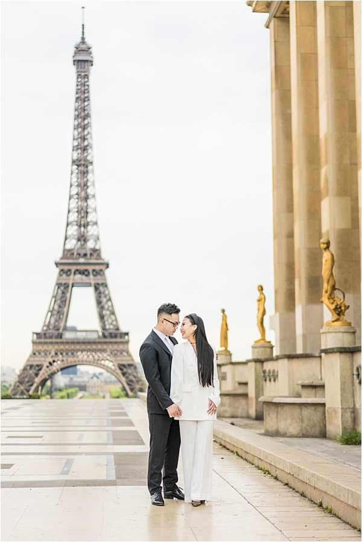 A couple poses together on the Trocadéro esplanade in Paris, with the Eiffel Tower centered directly behind them in the background. The man wears a black suit with a black bow tie, while the woman wears an all-white trouser suit with wide-leg pants; they are facing each other and holding hands. The gold-painted statues along the colonnade of the Palais de Chaillot are visible on the right side of the frame, adding architectural context to the outdoor urban setting. This is a full-length portrait shot with a clean, modern styling approach.
