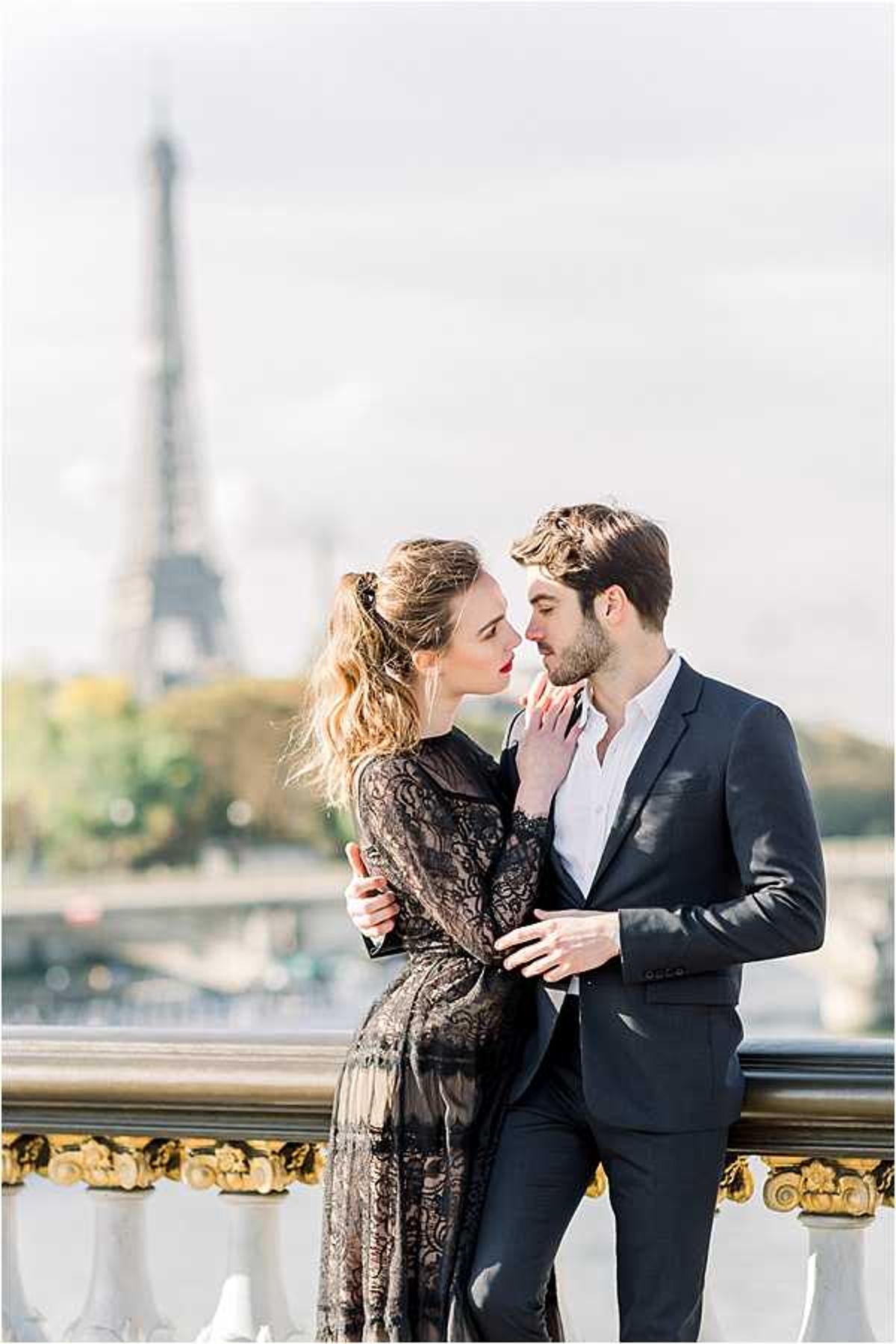 A couple portrait shot taken outdoors on the Pont Alexandre III bridge in Paris, with the Eiffel Tower softly out of focus in the background. The woman wears a long-sleeve black lace dress with a sheer overlay and has her hair pulled back loosely, wearing red lipstick; she is leaning in close to the man and touching his chin. The man is dressed in a dark navy suit jacket with a white open-collar shirt. The composition is a medium portrait framing the two subjects against the gold-detailed ornamental bridge railing, with the Seine River visible in the midground.