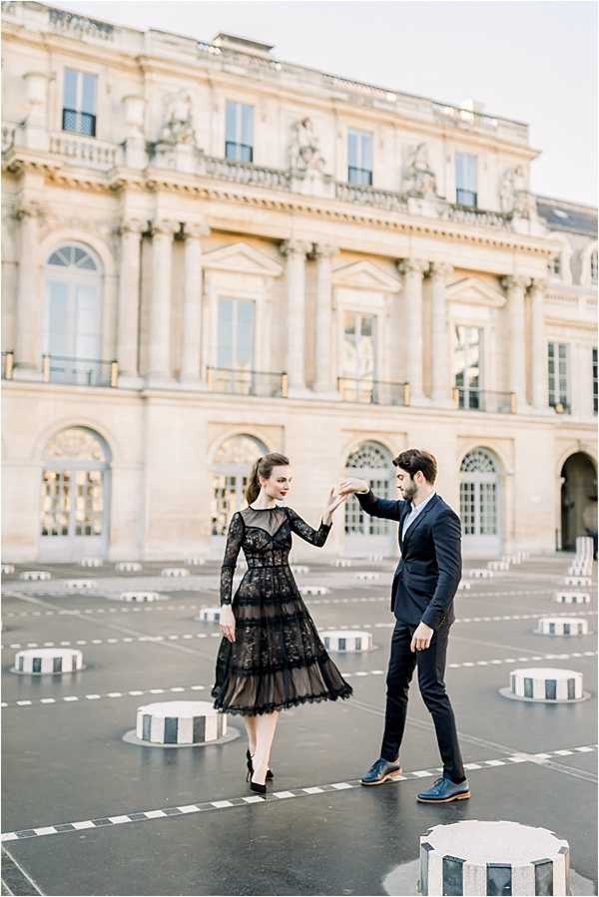 A couple poses in a dance-like stance at the Palais Royal courtyard in Paris, surrounded by the iconic black-and-white striped columns of the Buren installation. The woman wears a black lace midi dress with long sleeves, a tiered skirt, and black heels, while the man is dressed in a dark navy suit with blue leather oxford shoes. He is holding her hand aloft as if leading her in a spin, and both are looking at each other. The image is a wide portrait shot with the classical French neoclassical palace facade softly blurred in the background, giving the composition depth. The styling is polished and urban, with a classic Parisian aesthetic.