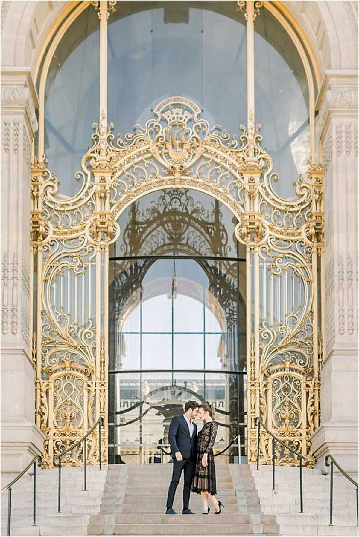 A couple shares an intimate forehead-to-forehead moment on the steps of a grand Beaux-Arts building featuring an ornate gold wrought-iron arched gate with intricate scrollwork and gilded detailing — consistent with the Petit Palais in Paris. The woman wears a black lace midi dress with sheer overlay and black heels, while the man is dressed in a navy suit. The composition is a full-length portrait shot framed centrally within the arch, emphasizing the scale of the architecture against the two figures. Potential venue feature image.