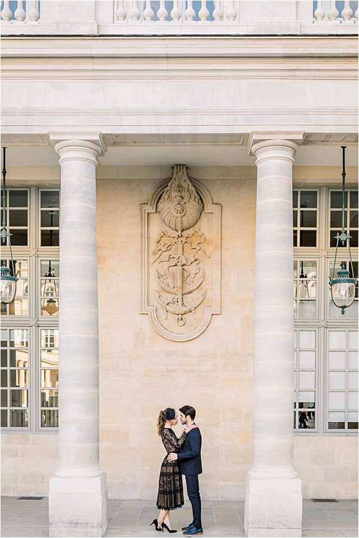 A couple shares a close embrace and near-kiss in front of a grand neoclassical building facade, likely the Palais Royal in Paris, featuring tall stone columns, ornate carved stone medallion relief, and vintage green patina lanterns flanking the archway. The woman wears a black lace midi dress with long sleeves and black heels, her hair styled up, while the man wears a navy suit. The shot is a wide portrait composition that emphasizes the scale of the pale limestone architecture against the small figures of the couple below. Potential venue feature image.