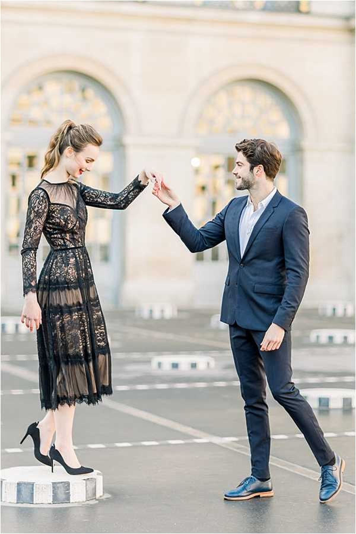 An engagement or couples portrait session in an outdoor urban plaza, likely in Paris, with a classical Haussmann-style stone building featuring arched windows visible in the background. The woman wears a long-sleeve black lace midi dress with a sheer skirt overlay and black high heels, standing on a low decorative bollard while the man holds her hand as if guiding her in a spin. The man is dressed in a fitted navy blue suit with a white shirt and blue leather oxford shoes. The pair are laughing and facing each other in a candid, playful moment. The image is a medium full-body shot with a shallow depth of field that softly blurs the architectural background.