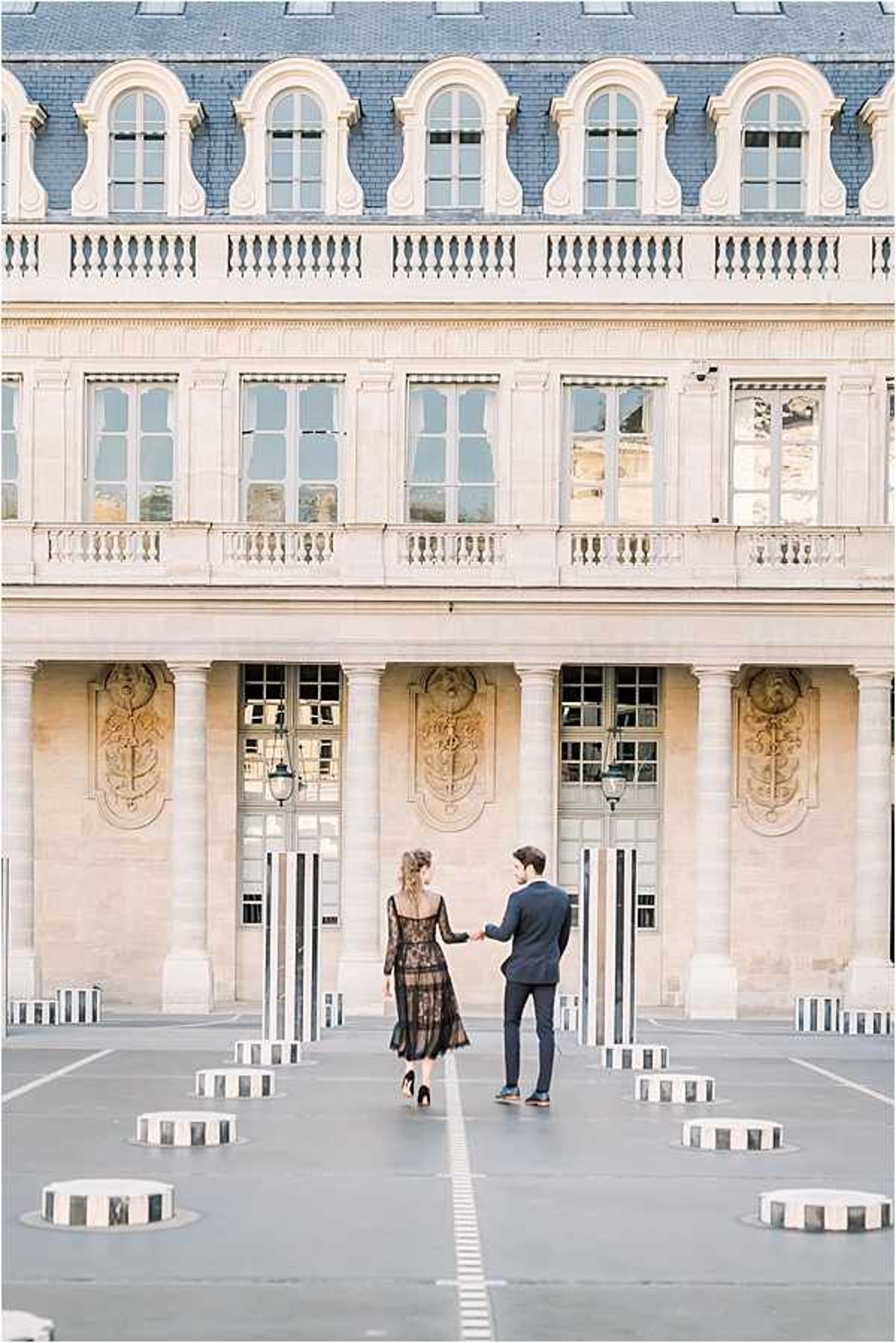 A couple portrait session shot outdoors at the Palais Royal courtyard in Paris, featuring the iconic black-and-white striped Colonnes de Buren installation in the foreground. The woman wears a black lace midi dress with long sleeves and black heels, while the man wears a fitted navy suit; they are holding hands and walking away from the camera toward the classical French architecture. The wide shot captures the full facade of the building with its arched windows, stone columns, ornate bas-relief panels, and wrought-iron lanterns. The composition is vertical and centered, emphasizing the architectural grandeur as a backdrop to the couple.