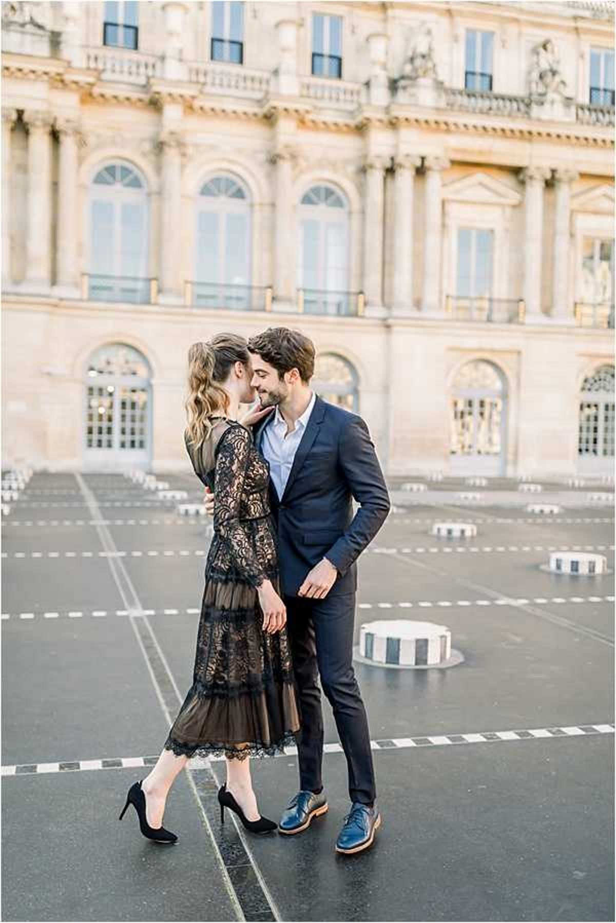 A couple shares an intimate moment outdoors in a courtyard in front of a large classical French stone building with arched windows, columns, and sculptural detailing, consistent with a Paris landmark. The woman wears a midi-length black lace dress with long sleeves and a sheer hem, paired with black suede stiletto heels; the man wears a navy suit with a light blue dress shirt and blue leather brogue shoes. They are facing each other closely, with the man's hand on her face, suggesting a near-kiss or tender exchange. The image is a full-length portrait shot with the building softly blurred in the background, captured in warm, diffused natural light.