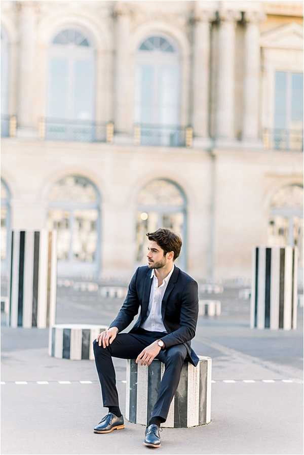 A solo portrait of a groom seated on one of the black-and-white striped columns of the Palais Royal courtyard in Paris, identifiable by Daniel Buren's Les Deux Plateaux installation visible in the background. The groom wears a navy suit with a white open-collar dress shirt and navy Oxford shoes, with a watch on his wrist. He is looking to one side in a relaxed, candid pose. The neoclassical architecture of the Palais Royal is softly blurred in the background. The shot is a medium full-length portrait taken at ground level with a shallow depth of field, captured in soft warm natural light.