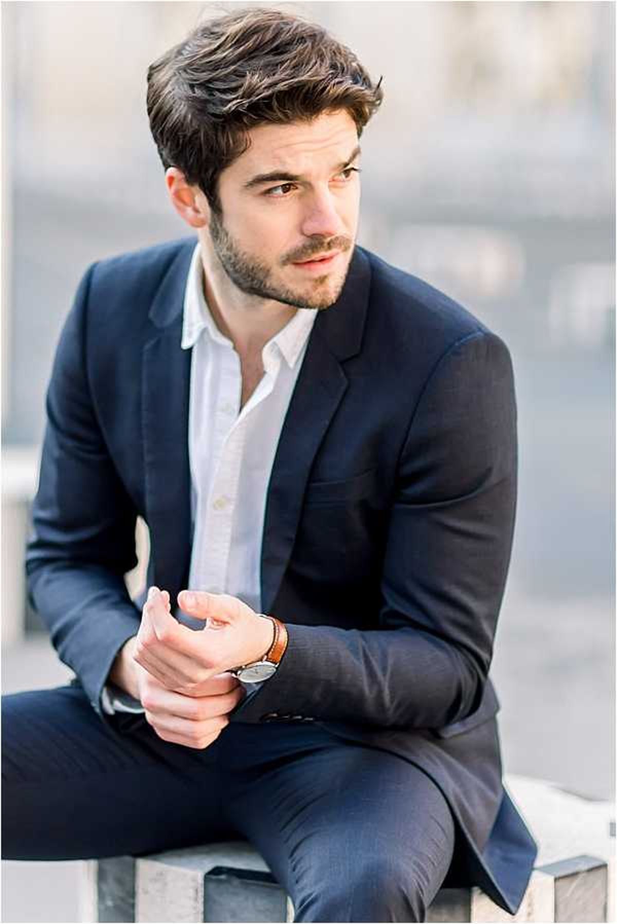 A close-up portrait of a groom seated outdoors, wearing a navy suit jacket, an open-collar white dress shirt with no tie, navy trousers, a brown leather bracelet, and a minimalist watch. He is looking slightly off-camera to one side with his hands clasped in front of him. The background is heavily blurred, suggesting an urban outdoor setting with light-toned surfaces. The styling is relaxed and modern, with the open collar and casual accessories giving a smart-casual aesthetic. The image is shot in natural light with a shallow depth of field.