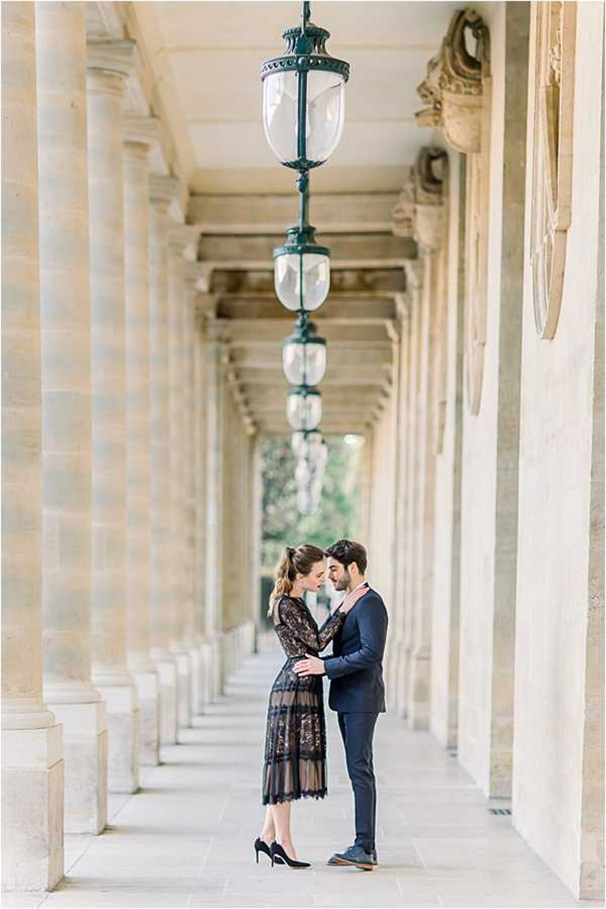 A couple shares an intimate moment standing beneath a row of ornate dark green hanging lanterns in a classical Parisian arcade, likely the Palais-Royal. The woman wears a long-sleeve black lace midi dress with black heels, her hair pulled back in a low ponytail, while the man wears a fitted navy suit with a light pink tie. They are facing each other closely with hands intertwined, photographed in a full-length portrait composition with the receding colonnade creating strong architectural symmetry and depth behind them. Potential venue feature image.