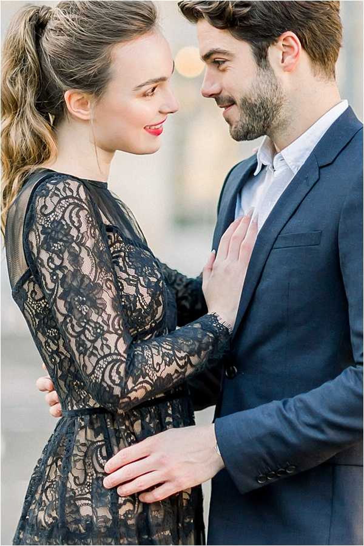 A couple portrait shot in a close-up composition, with the two facing each other in an intimate pose. The woman wears a long-sleeve black lace dress over a nude lining, her dark hair pulled back in a high ponytail, and she has red lipstick; the man wears a navy blue suit with a light blue open-collar shirt. She has one hand placed on his chest while he holds her waist. The background is softly blurred with a warm bokeh light visible, suggesting an outdoor urban setting, likely Paris. The styling is classic and polished with a dark, graphic color palette.