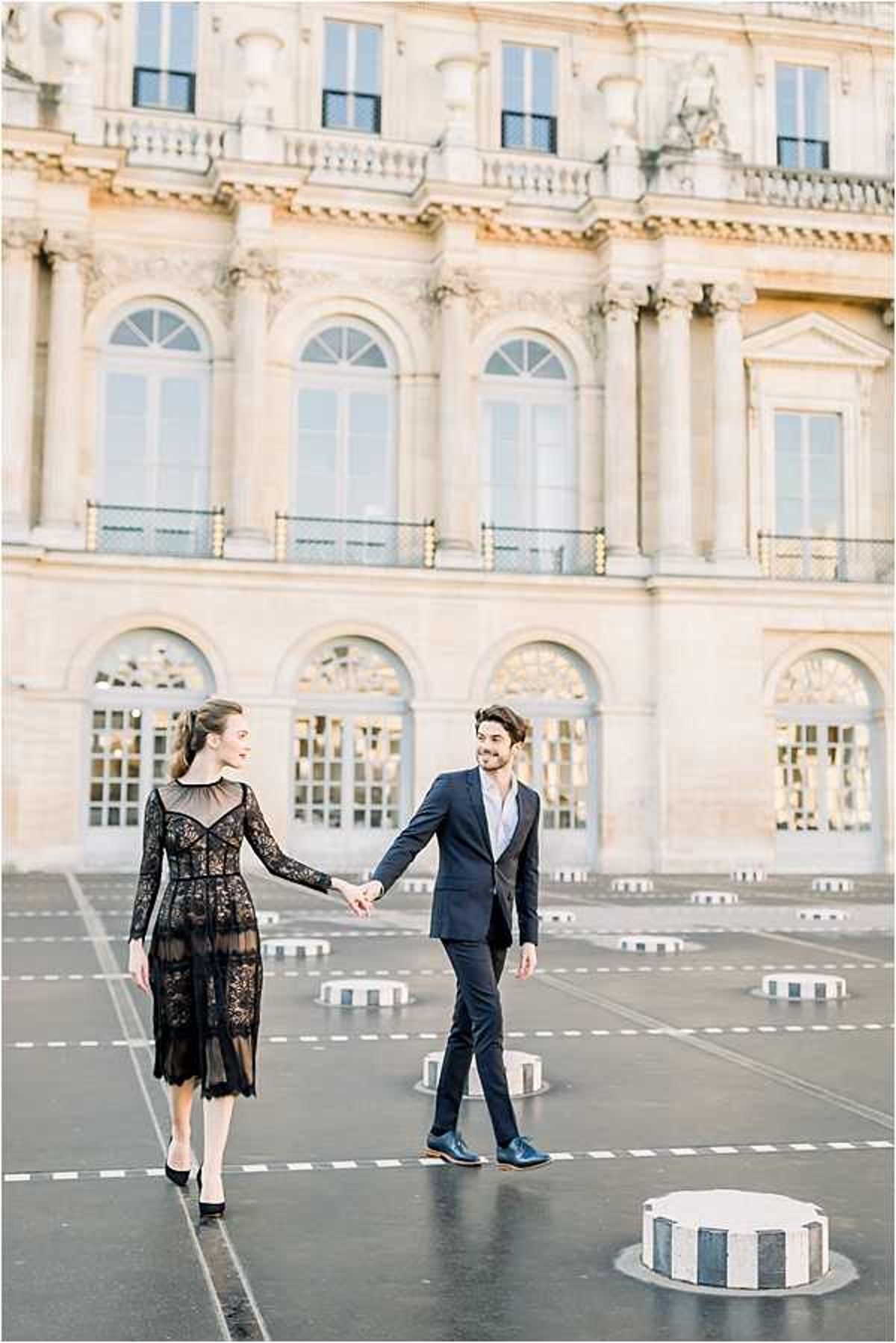 A couple poses for an engagement or pre-wedding portrait session outdoors in a Parisian courtyard, identifiable by the grand Neoclassical limestone facade with arched windows and ornate detailing in the background, consistent with the Palais Royal or Louvre area. The woman wears a black midi-length lace dress with long sleeves and a sheer overlay, paired with black heels, while the man wears a fitted navy suit with a light blue dress shirt and navy leather shoes. They are holding hands and walking toward each other, both smiling, with striped cylindrical bollards visible across the open paved courtyard. Wide portrait shot with soft, warm natural light and a bright, airy tonal palette. Potential venue feature image.