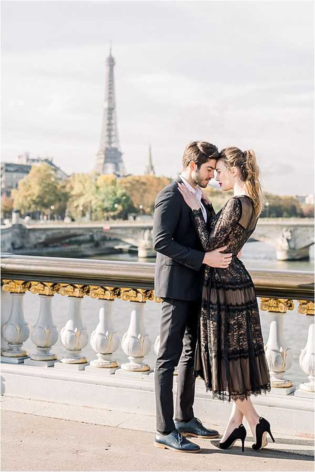 A couple shares an intimate moment on the Pont Alexandre III bridge in Paris, with the Eiffel Tower and the Seine River visible in the background. The woman wears a black long-sleeve lace midi dress with sheer tiered panels and black heeled pumps, with her hair pulled back in a loose updo, while the man wears a dark navy suit with oxford shoes. They are embracing closely, foreheads nearly touching, in a classic portrait composition with the bridge's white stone balustrade and gold-gilded railings framing the scene. The overall styling is polished and Parisian, with a dark, moody color palette against the soft autumn light.