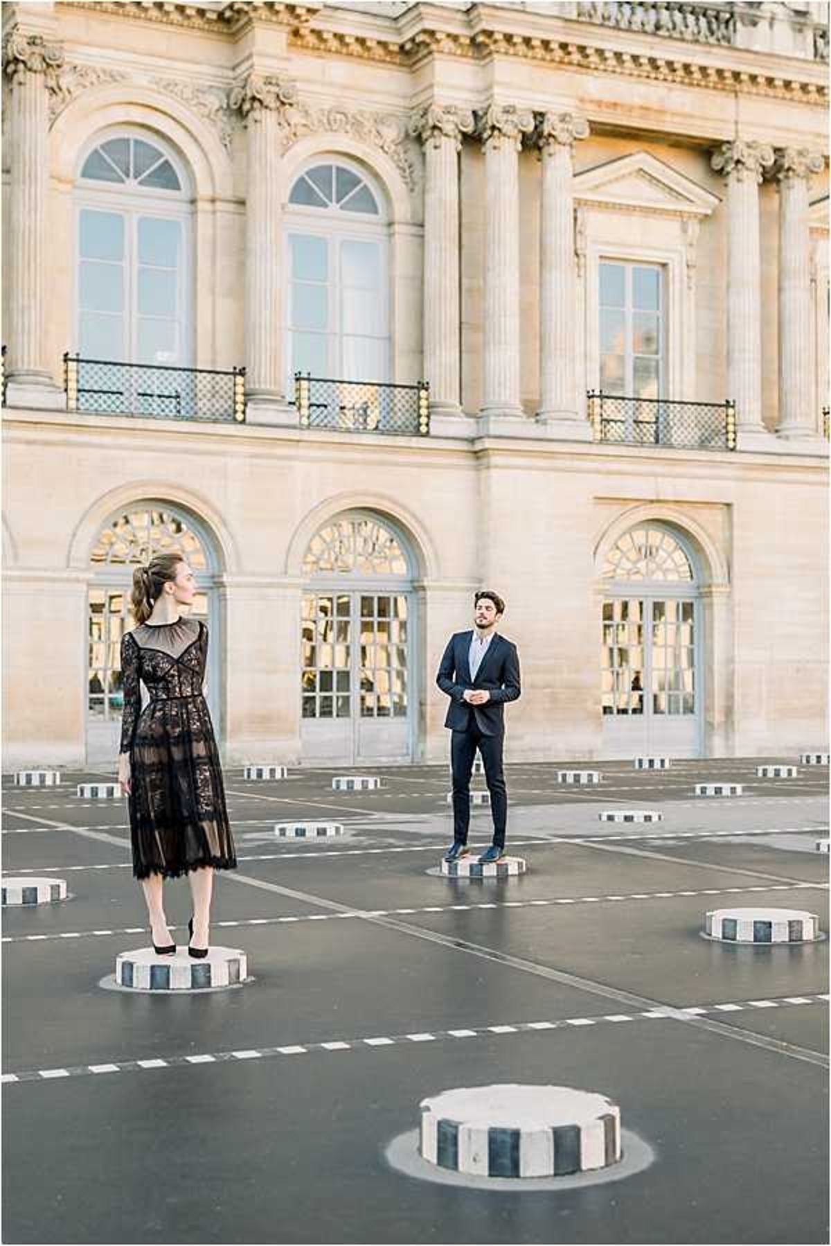 A couple poses outdoors in the courtyard of the Palais Royal in Paris, standing apart on the signature striped black-and-white cylindrical columns of the Daniel Buren installation. The woman wears a midi-length black lace dress with sheer overlay panels and long sleeves, paired with black stiletto heels and her hair styled up; the man wears a fitted dark navy suit with a light-colored shirt. The wide portrait shot captures the classical French limestone architecture — arched windows, Corinthian columns, and wrought-iron balconies — as a backdrop. The styling is modern and editorial with a monochrome palette. Potential venue feature image.