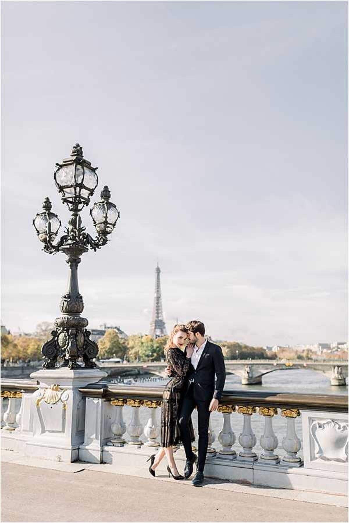 A couple poses together on the Pont Alexandre III bridge in Paris, with the Eiffel Tower visible in the background across the Seine River. The woman wears a black lace midi dress with black stiletto heels, while the man is dressed in a dark navy suit; he leans in to kiss her cheek as she tilts her head toward him. The bridge's ornate white and gold balustrade and a large decorative iron lamp post are prominent in the foreground, framing the couple. This is a wide portrait shot taken in natural daylight with a bright, airy exposure.