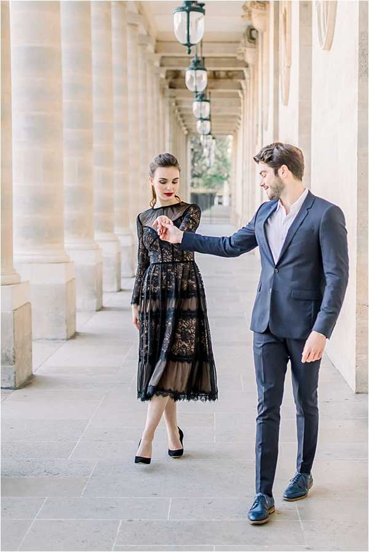 A couple poses in a covered outdoor colonnade, likely at the Palais-Royal in Paris, with a row of stone columns and pendant lanterns receding into the background. The man, dressed in a navy blue suit with a light blue open-collar shirt and navy leather oxford shoes, is gently holding the woman's hand as she walks toward him. The woman wears a black lace midi dress with long sleeves, a sheer overlay with floral lace paneling, and black pointed-toe heels, with her dark hair pulled back and bold red lipstick. The shot is a full-length portrait taken in natural light with a shallow depth of field that blurs the colonnade behind them.