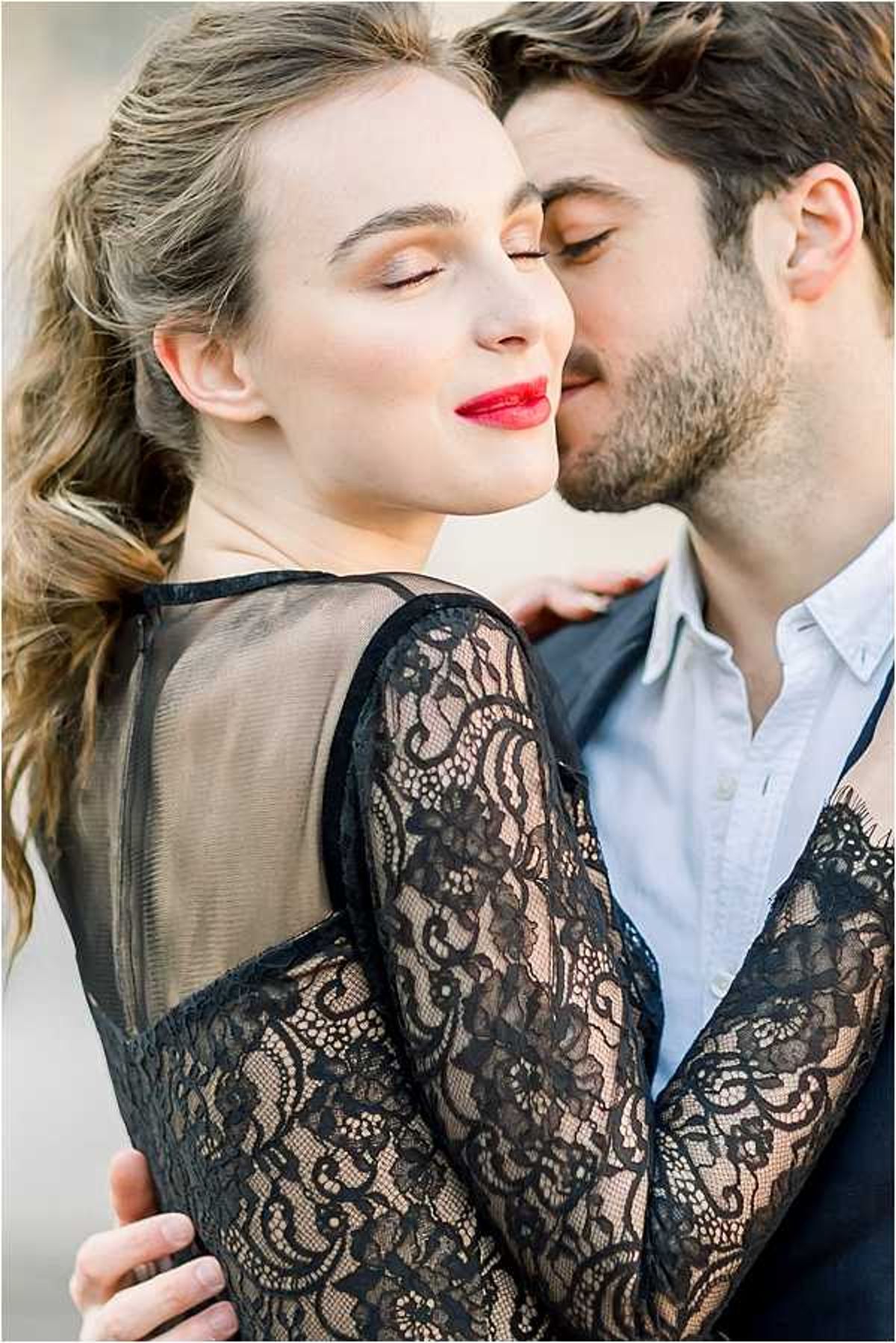 A close-up portrait of a couple in an intimate pose, with the man kissing the woman's cheek while she closes her eyes. The woman wears a black lace long-sleeve dress with sheer panels and floral lace detailing, paired with bold red lipstick and warm eye makeup; her wavy hair is loosely styled. The man wears a light blue dress shirt under a dark blazer. The shot is tightly cropped, focusing on their faces and the upper body, with a soft blurred background. The overall styling is modern and fashion-forward with a monochromatic black outfit contrasted by the red lip.