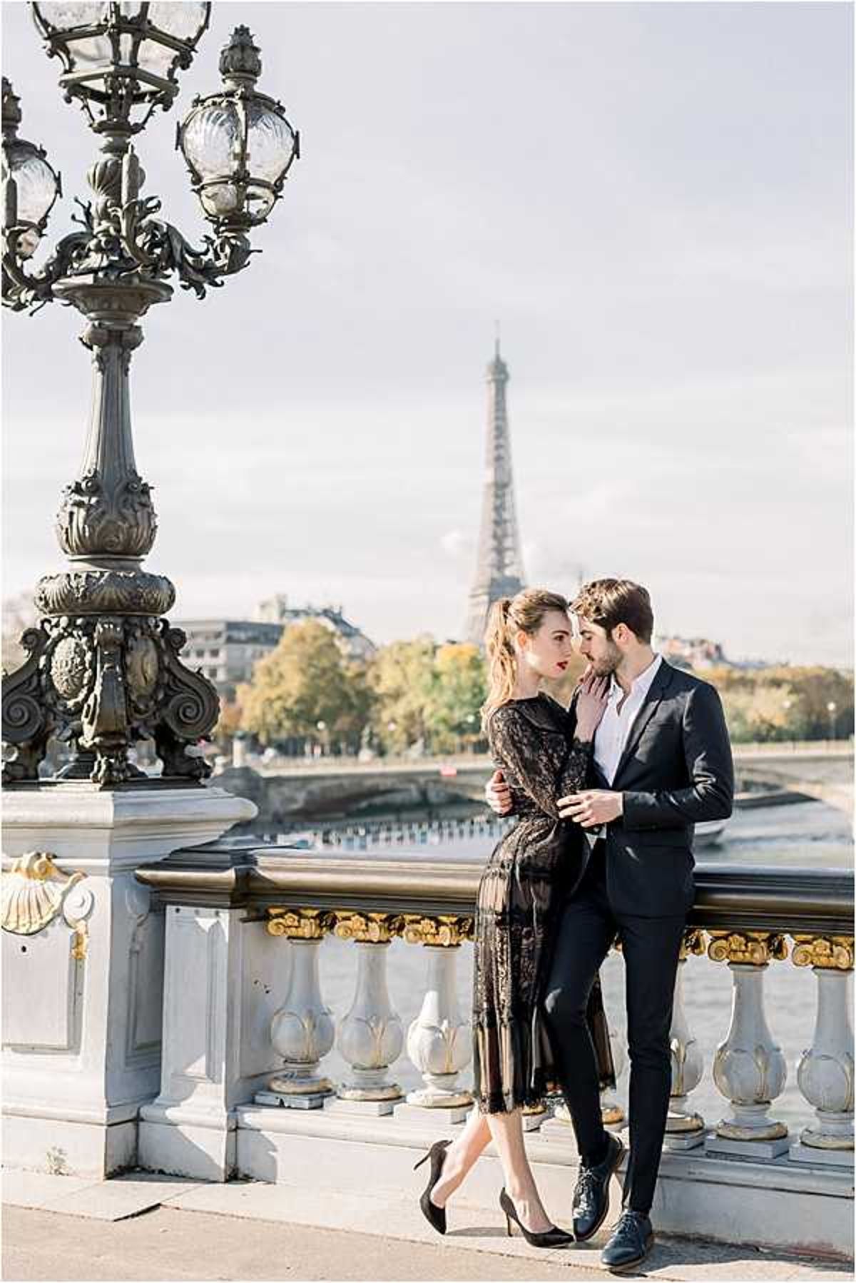 A couple poses together on the Pont Alexandre III bridge in Paris, with the Eiffel Tower visible in the background. The woman wears a black lace midi dress with long sleeves and black stiletto heels, while the man wears a dark navy suit with an open-collar white shirt and oxford shoes. The man leans in close to the woman's face as she looks toward the camera, with his hand resting on her cheek in an intimate gesture. The shot is a medium portrait taken in natural daylight, framed by the bridge's ornate gilt and stone balustrade and one of its signature bronze candelabra lamp posts on the left.