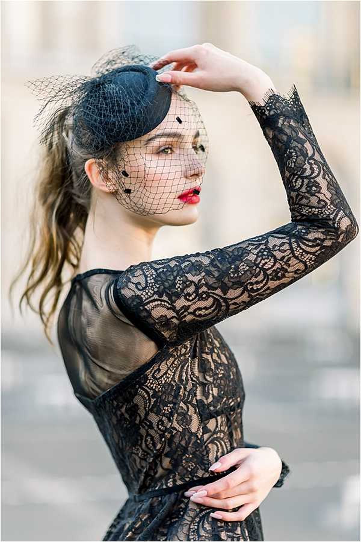 A close-up portrait of a bride wearing a black lace long-sleeve dress with a fitted bodice and sheer mesh panels, paired with a black birdcage veil fascinator hat adorned with small polka dots. She has bold red lipstick, a loose ponytail, and light pink nail polish, with one hand raised to hold the fascinator. The background is blurred but suggests an outdoor urban or architectural setting. The styling is classic and fashion-forward with a monochromatic black bridal look.