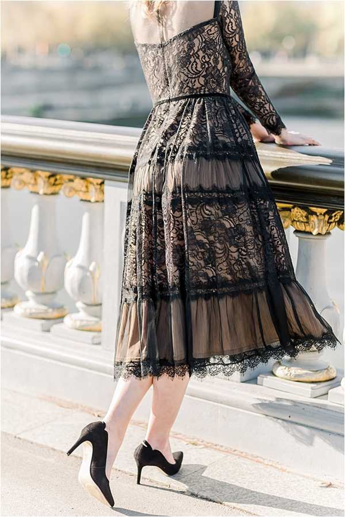 A close-up portrait shot taken from behind, focusing on a woman wearing a black lace overlay dress with a nude/champagne underlining, featuring long lace sleeves, a fitted bodice, and a tiered midi-length skirt with scalloped lace hem detail. She is wearing black satin pointed-toe stiletto heels and is resting her hand on an ornate railing with white and gold-gilded balustrade details, consistent with Parisian bridge architecture such as the Pont Alexandre III. The composition highlights the intricate layered lace construction of the dress as a key style detail. The overall styling aesthetic is classic Parisian with a dark, formal color palette.