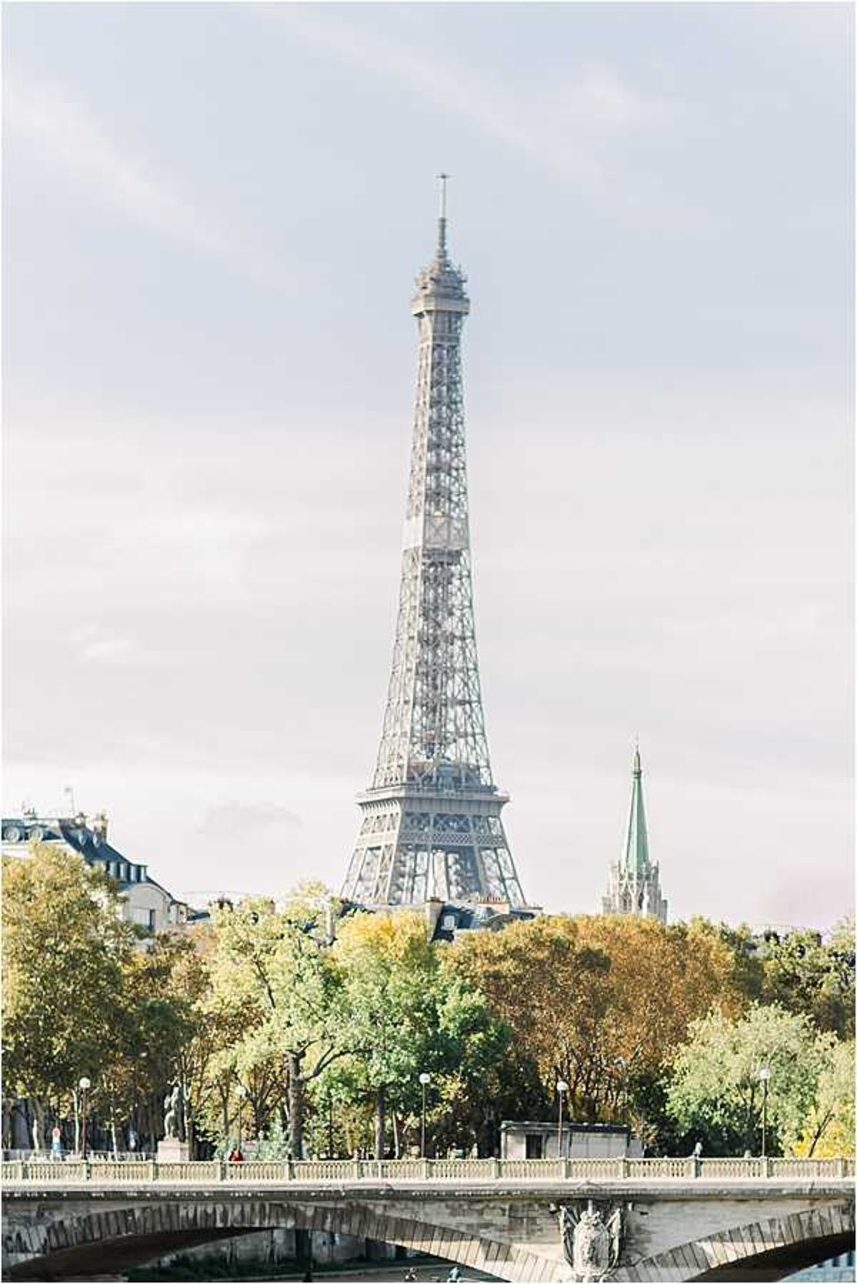 A wide-angle location shot taken in Paris, France, showing the Eiffel Tower rising above the treeline along the Seine river, with a stone bridge visible in the foreground and Haussmann-style buildings and a church spire to the sides. No people, couple, or wedding-specific elements are visible in the frame. The image appears to be a venue or location establishing shot, likely used to set the Paris context for a wedding editorial. Potential venue feature image.