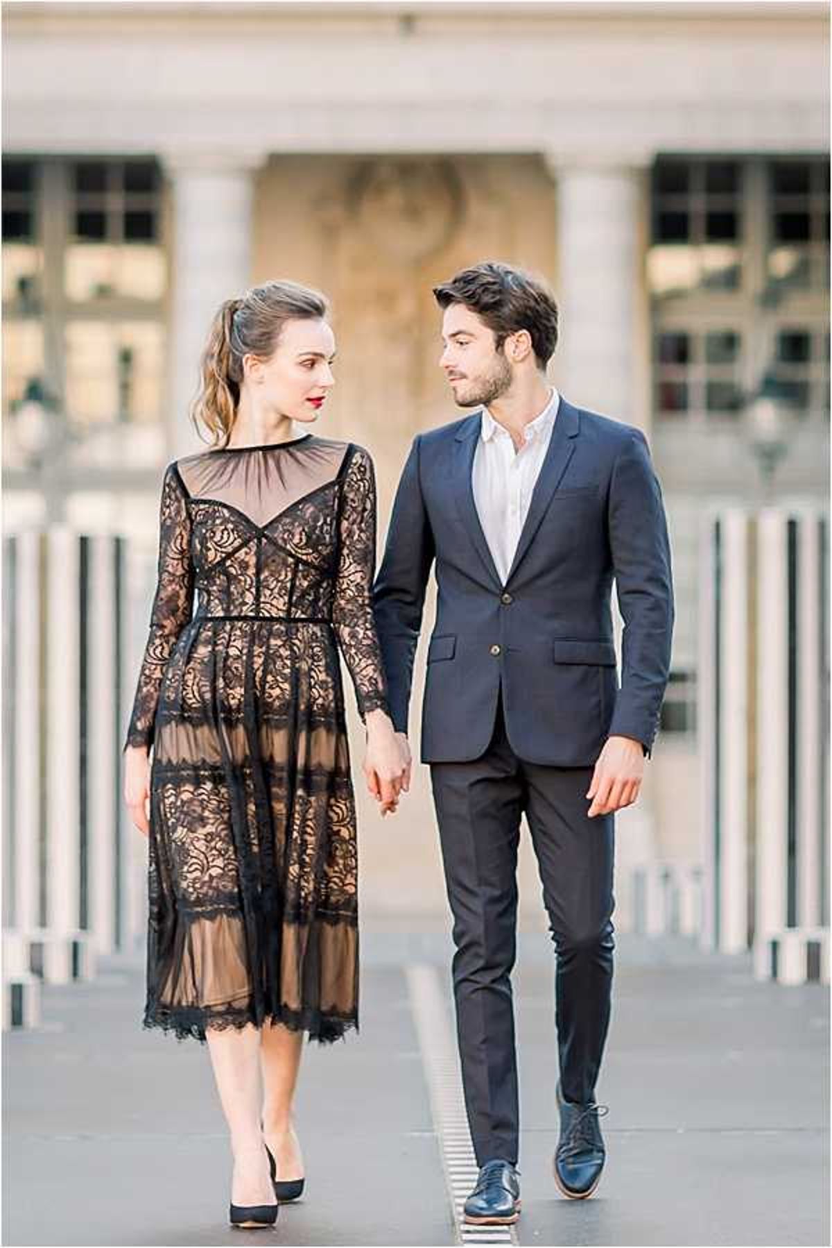 A couple portrait shot outdoors in front of a classical Parisian building with columns and large windows visible in the soft-focus background, likely the Palais Royal. The woman wears a midi-length black lace overlay dress with long sleeves, a sweetheart neckline visible beneath the sheer bodice, and black heels, styled with red lipstick and her hair loosely pinned up. The man wears a fitted navy suit jacket with dark trousers and a white open-collar shirt, finished with dark oxford shoes. The two are walking toward the camera, holding hands and looking at each other, shot as a mid-length portrait with a shallow depth of field that keeps focus on the couple.