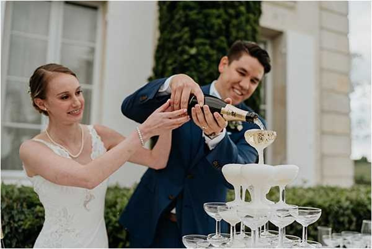 The bride and groom are pouring champagne together into a coupe glass tower during an outdoor reception, with champagne visibly overflowing down the pyramid of glasses. The bride wears a white lace dress with a pearl necklace and has her hair pulled back, while the groom wears a navy blue suit with a white boutonnière. Both are smiling as they hold the bottle together. The setting appears to be the grounds of a French chateau or manor house, visible in the soft-focus background. The shot is a medium close-up portrait capturing the couple from the waist up alongside the champagne tower.