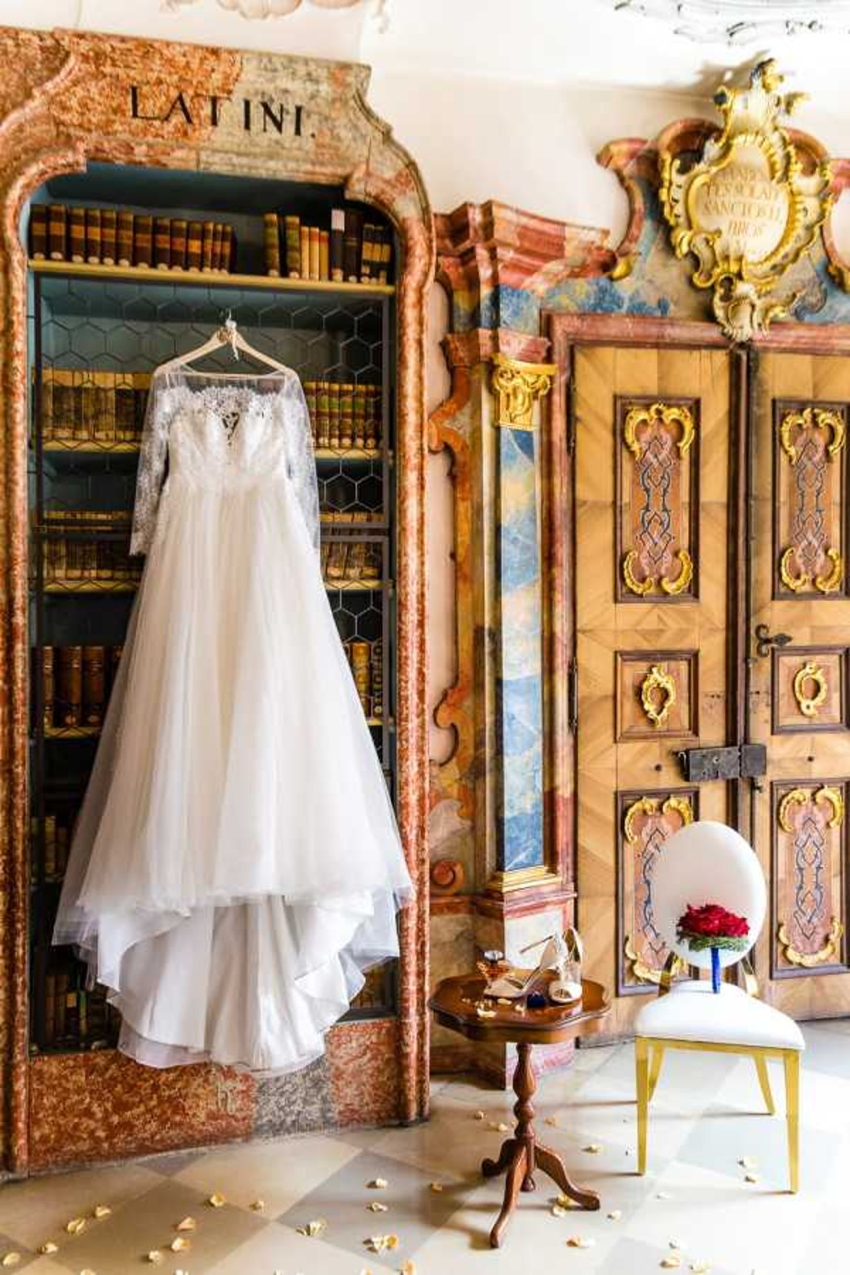 A bridal flat-lay style getting-ready detail shot taken indoors in what appears to be a historic baroque library or palace room. The ivory wedding gown — a full A-line silhouette with long lace sleeves, a lace illusion bodice, and layered tulle skirt — hangs on a hanger in front of an ornate bookcase labeled 'LATINI' filled with antique leather-bound books, framed by decorative carved and marbled surrounds. To the right, a small wooden pedestal table holds gold strappy bridal heels and accessories, while a white and gold upholstered chair displays a compact bouquet of deep red roses with a cobalt blue ribbon wrap. The room features elaborate gilded baroque doors with inlaid wood panels and painted decorative columns, and scattered flower petals are visible on the marble floor. The overall styling is classic and formal, set within a richly decorated historic interior.