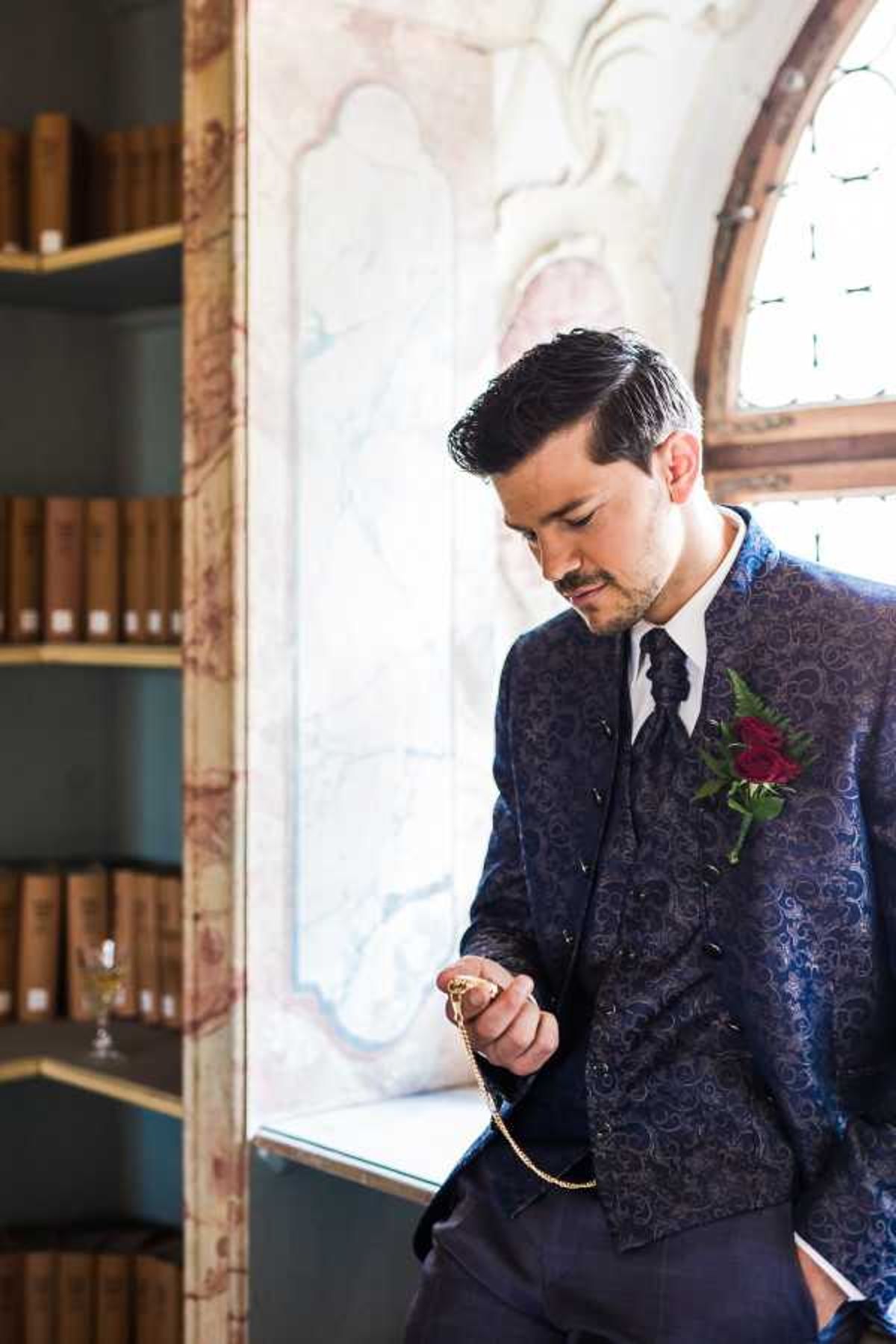 A groom portrait taken indoors, likely inside a chateau or historic venue library, with ornate plasterwork and an arched stained-glass window visible in the background. The groom is wearing a navy blue paisley-patterned suit jacket and matching waistcoat, a white dress shirt, and a dark navy knot tie, with a deep red rose and fern boutonniere on his lapel. He is looking downward at a gold pocket watch and chain held in his hand, posed leaning against the window ledge. Antique leather-bound books on dark shelving are visible to the left, along with a small wine glass, adding to the classic, library setting of the portrait.