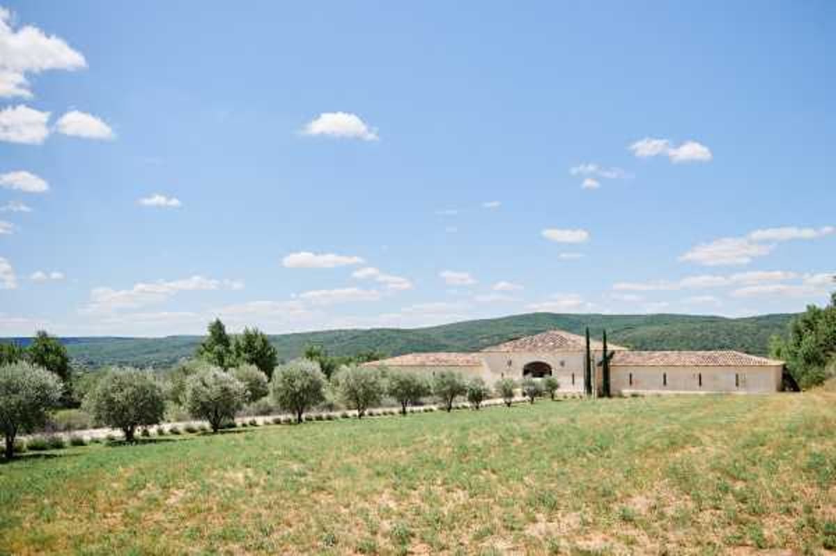 Wide exterior shot of a low-profile stone venue building with a terracotta-tiled roof and arched entrance, set within a rural Provençal landscape. The property features a row of olive trees lining what appears to be a driveway, with open fields in the foreground and tree-covered hills in the background. No people are visible in the frame. The image is taken from a distance, showcasing the full scale and setting of the venue. Potential venue feature image.