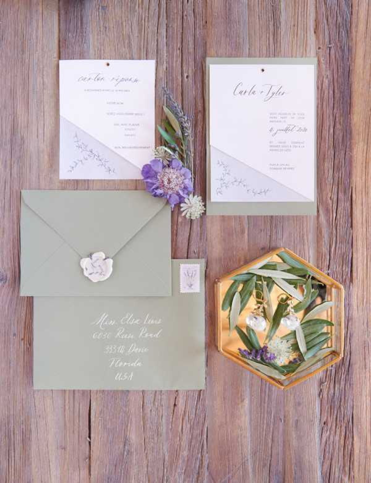 A flat lay detail shot of a wedding stationery suite arranged on a weathered wood surface. The suite includes a sage green envelope with white calligraphy addressing and a white wax seal, a menu card, and an invitation card for 'Carla + Tyler' dated 6 juillet 2018, both printed on white card stock with a sage green diagonal color block design and delicate botanical line illustrations. A small cluster of dried purple scabiosa flowers and olive or eucalyptus sprigs is tucked between the cards. A gold hexagonal glass tray sits to the right, holding olive foliage, lavender sprigs, and a pair of delicate gold drop earrings with clear crystal pendants. The color palette is sage green, white, and soft purple with gold accents, giving the styling a botanical, garden-inspired feel.