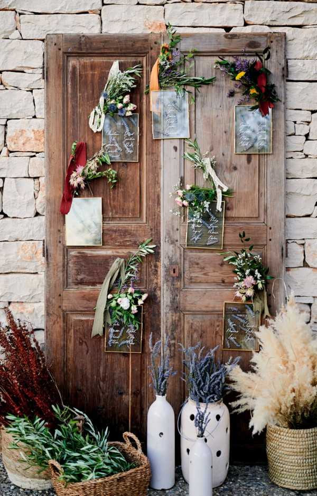 A close-up detail shot of a rustic wedding seating chart display mounted on a pair of aged wooden double doors leaning against a stone wall. Multiple gold-framed acrylic or vellum seating chart cards with white calligraphy are attached across both door panels, each decorated with small mixed floral clusters featuring pink dahlias, lavender, olive branches, wildflowers, and tied with burgundy, sage green, and cream ribbons. At the base of the doors, an arrangement of decorative vessels is grouped together, including tall white ceramic vases with black dot detailing holding dried lavender and pampas grass, a wicker basket with olive branches, and a woven basket with dried reddish grasses. The overall styling is bohemian-rustic with a warm, earthy palette incorporating burgundy, sage, dusty pink, and natural dried botanicals.