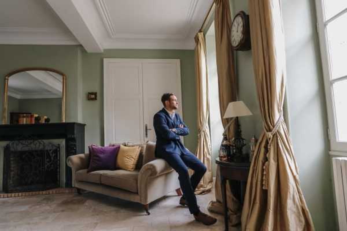 A groom in a navy blue suit with arms crossed sits on the arm of a beige velvet sofa, gazing out of a tall window with gold floor-length curtains. The interior room features sage green walls, ornate white crown molding, a fireplace with a gilt-framed mirror, and a small antique side table with a lamp and glassware. The setting is an indoor salon or sitting room, likely within a chateau or manor house. This is a medium-wide portrait shot taken during the getting-ready or pre-ceremony period.