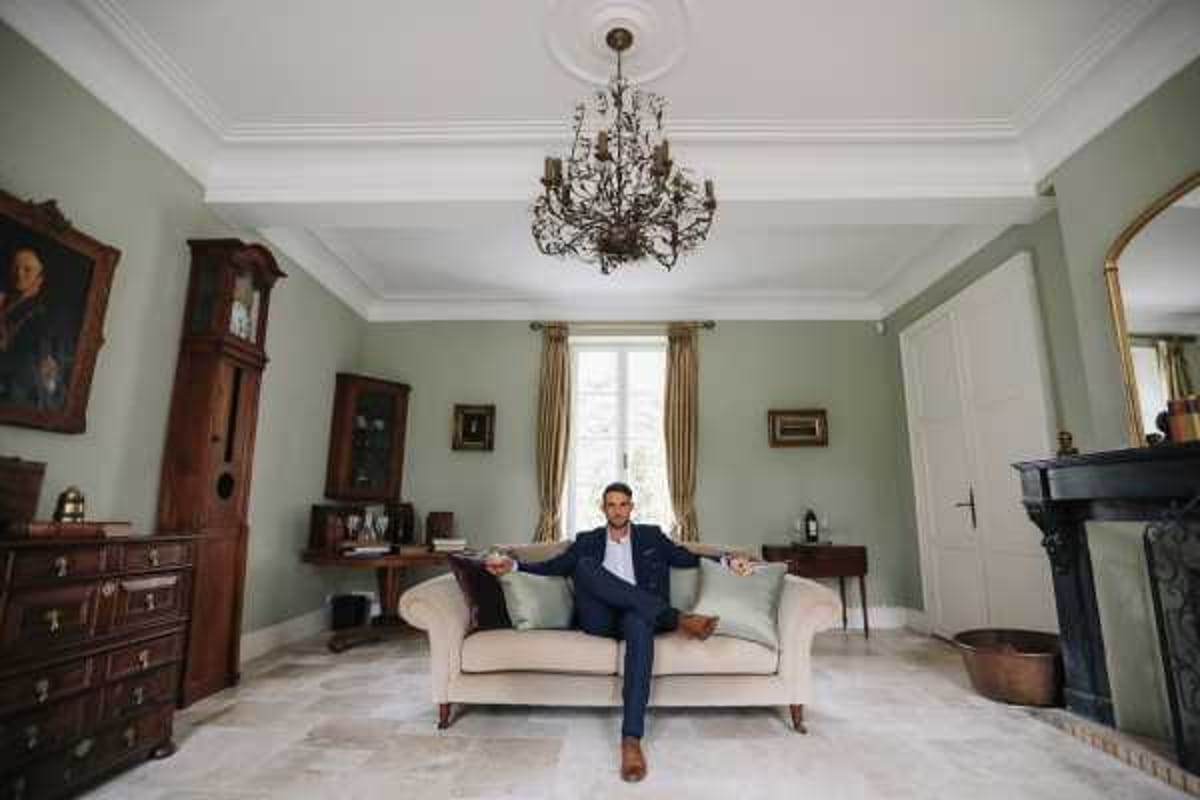 A groom in a navy blue suit with a white dress shirt sits relaxed on a cream chaise longue in a formal interior room, arms spread wide across the back of the sofa. The room features sage green walls, ornate white crown molding, a black marble fireplace, a bronze branch-style chandelier, gold-framed mirror, dark wood antique furniture including a grandfather clock and chest of drawers, gold drape curtains, and marble tile flooring. Wide shot taken from a low, centered angle, placing the groom symmetrically within the room. Potential venue feature image.