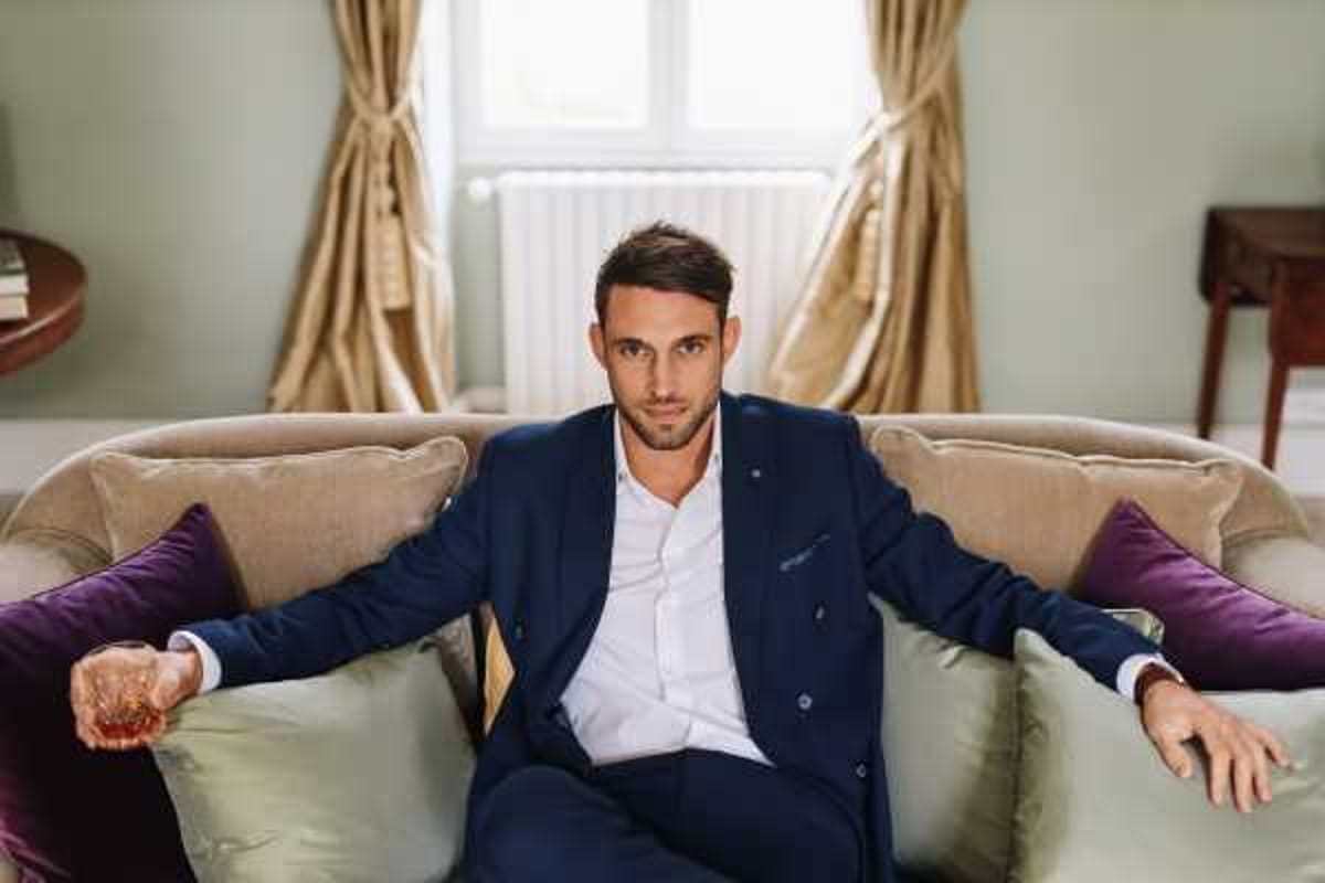 A groom getting-ready portrait taken indoors, likely in a hotel or chateau suite. The groom is seated on a velvet sofa with sage green and deep purple cushions, wearing a navy double-breasted suit jacket over a white dress shirt with no tie, holding a glass of whisky in his right hand. The room features gold curtains, a pale green wall, and natural window light from behind. The composition is a medium portrait shot with the subject facing the camera directly.
