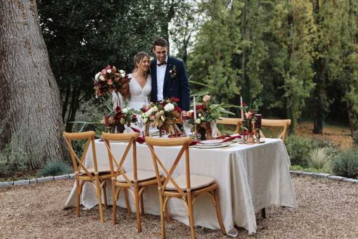 A couple stands together at a styled sweetheart table set outdoors on a gravel surface surrounded by trees and garden landscaping. The bride wears a fitted white V-neck gown and holds a large bridal bouquet featuring deep burgundy, coral, and cream blooms with tropical greenery and palm leaves, while the groom wears a navy double-breasted suit with a bow tie. The rectangular table is dressed with a cream linen tablecloth and set with natural wood cross-back chairs, gold candlestick holders with red taper candles, and floral arrangements in burgundy, coral, and ivory with lush greenery echoing the bouquet palette. The overall styling is earthy and moody with a warm autumn feel, combining rustic and bohemian design elements. Wide portrait shot capturing both the couple and the full table setup.