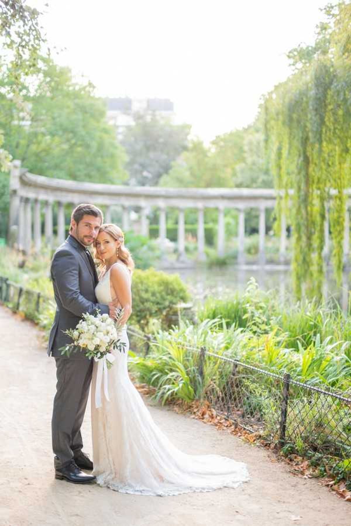 A couple portrait taken outdoors in a formal French park, with the groom standing behind the bride and embracing her as both face the camera. The bride wears a fitted ivory lace gown with a train and holds a bouquet of white roses and greenery with trailing ivory ribbons; the groom wears a charcoal grey suit with a dark tie. The background features a stone balustrade colonnade beside a small pond, with a weeping willow at right and lush garden plantings enclosed by a black iron fence. The shot is a full-length portrait with soft, warm natural light, taken along a gravel path — likely in the Parc Monceau in Paris.