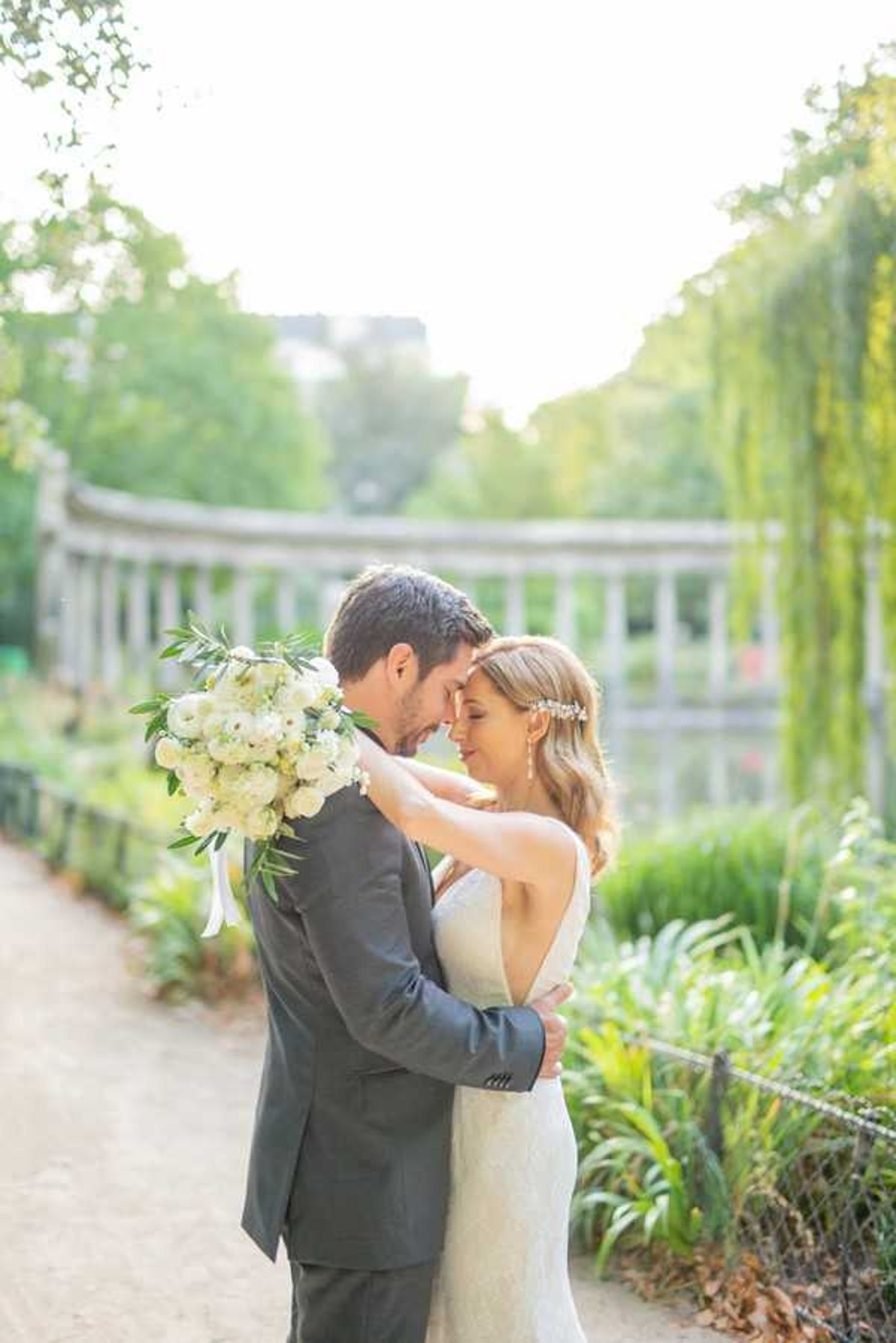 A couple portrait taken outdoors in a formal garden setting, with the bride and groom standing close together with foreheads nearly touching in an intimate pose. The groom wears a charcoal grey suit, while the bride wears a fitted ivory lace gown with a low back and a small floral hair accessory in her loose waves. She holds a lush bouquet of ivory ranunculus, white hydrangea, and greenery with trailing white ribbon. The background features a classical stone balustrade bridge and weeping willow foliage, softly blurred with warm golden-hour light. The shot is a medium portrait framed from the waist up.