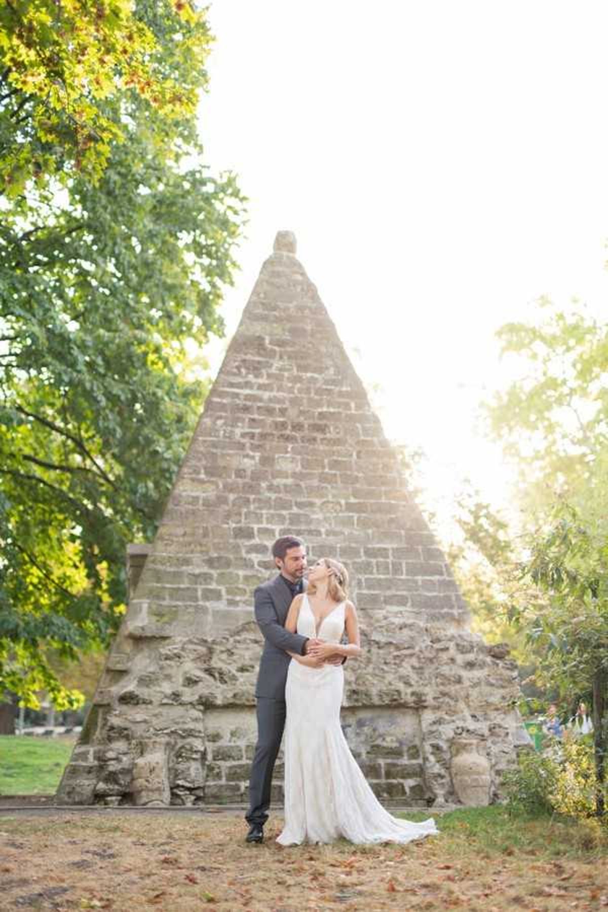 A couple portrait taken outdoors in a garden setting, with a large stone pyramid structure serving as the backdrop. The groom, wearing a charcoal grey suit, stands behind the bride with his arms wrapped around her waist, and the two are looking at each other. The bride wears a fitted, low-cut white gown with a short train and has her hair loosely styled with a floral hair accessory. The shot is a full-length portrait taken in warm golden-hour light, with the pyramid's stone masonry clearly visible behind the couple.