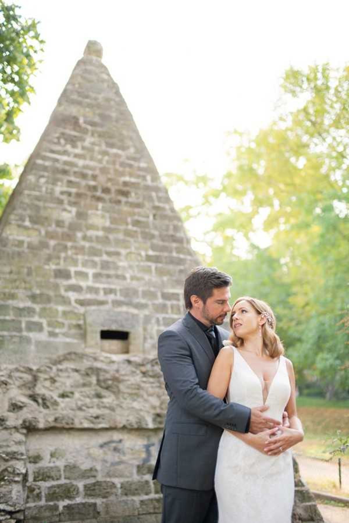 A couple portrait taken outdoors in warm, golden natural light. The groom, wearing a charcoal grey suit with a dark tie and a white boutonniere, stands behind the bride with his arms wrapped around her waist. The bride wears a fitted white gown with a deep V-neckline and thin straps, and her hair is styled in a loose updo with a small floral hair accessory. She looks up at the groom while he leans in close to her. The background features a distinctive pyramid-shaped stone structure built from large cut limestone blocks, surrounded by mature trees. The composition is a mid-length couple portrait with the stone pyramid centered in the background.