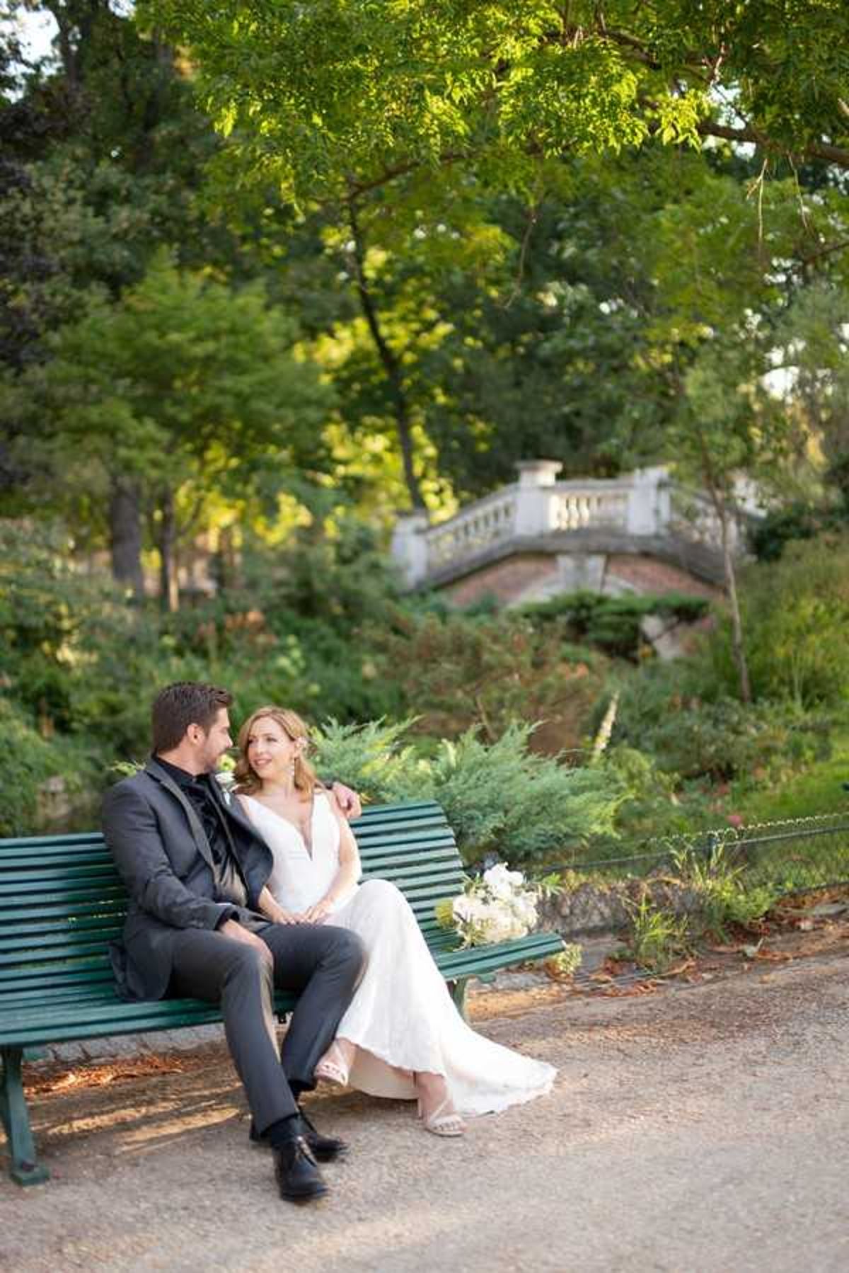 A couple portrait taken outdoors in a formal garden setting, with the bride and groom seated together on a dark green park bench. The groom wears a charcoal suit with a black tie, while the bride wears a fitted ivory sleeveless gown with a deep V-neckline; a white and cream bouquet rests on the bench beside her. The two are looking at each other and smiling in a relaxed, candid moment. In the background, a white stone ornamental bridge with balustrade railing is visible among lush greenery, suggesting a classic French public garden such as a Parisian park. The shot is a medium-wide portrait with warm natural light filtering through the tree canopy above.