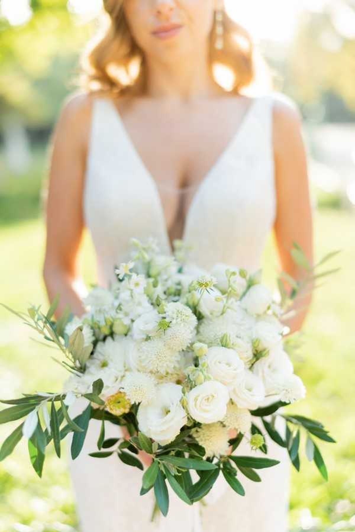 A bridal portrait detail shot taken outdoors, with the bride's face softly out of focus in the background while the bouquet is the primary subject in sharp focus. The bride wears a white fitted gown with a deep V-neckline and gold drop earrings, with her blonde hair loosely waved. The large, lush bouquet is composed of ivory garden roses, white lisianthus, white scabiosa, small white spray flowers, and green scabiosa buds, accented with trailing olive branch foliage, creating an organic, garden-style arrangement in an all-white and green palette.